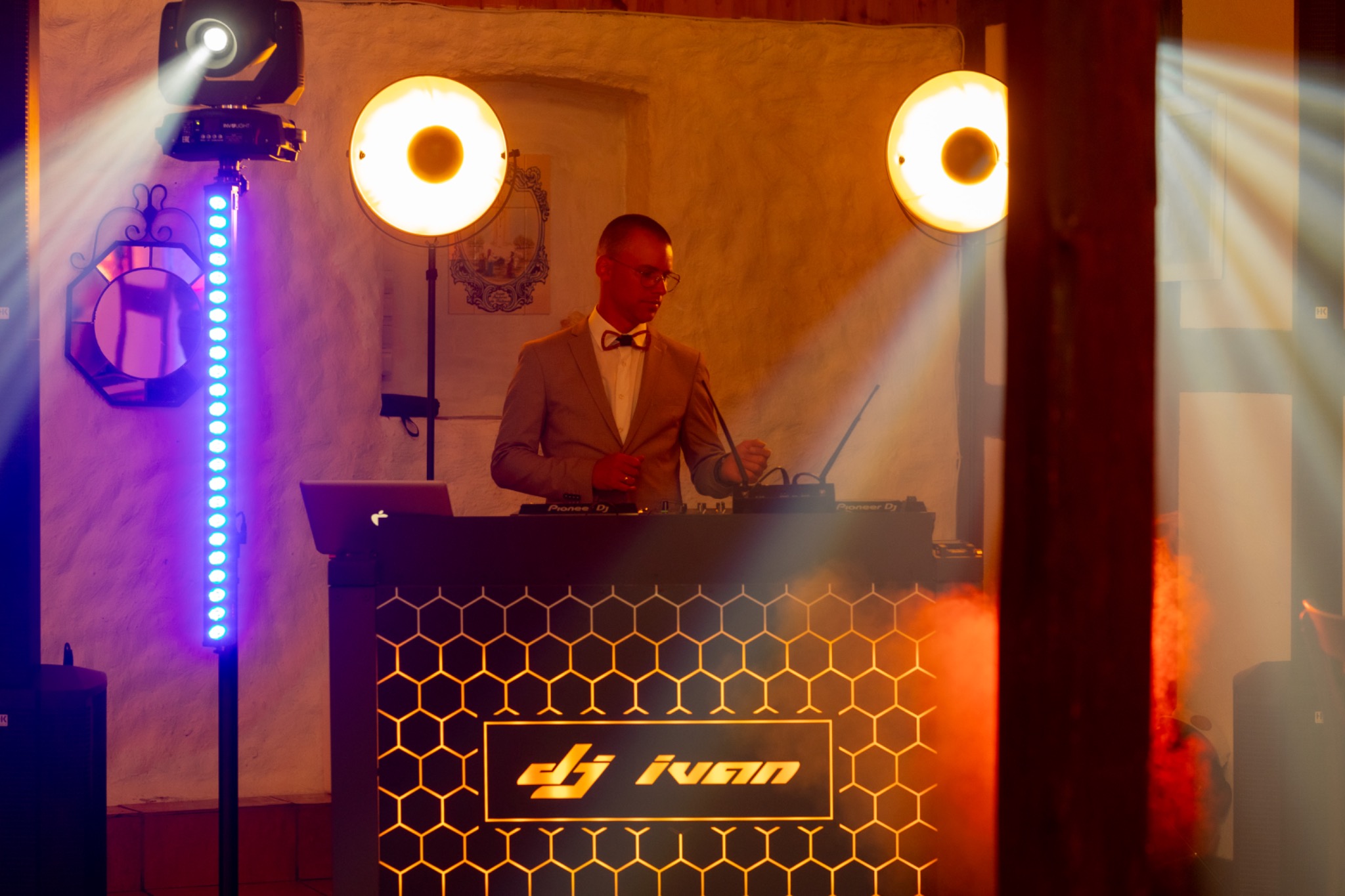DJ Ivan w trakcie występu, oświetlenie sceniczne z reflektorami i listwą LED, konsoleta z logo na tle ceglanej ściany.