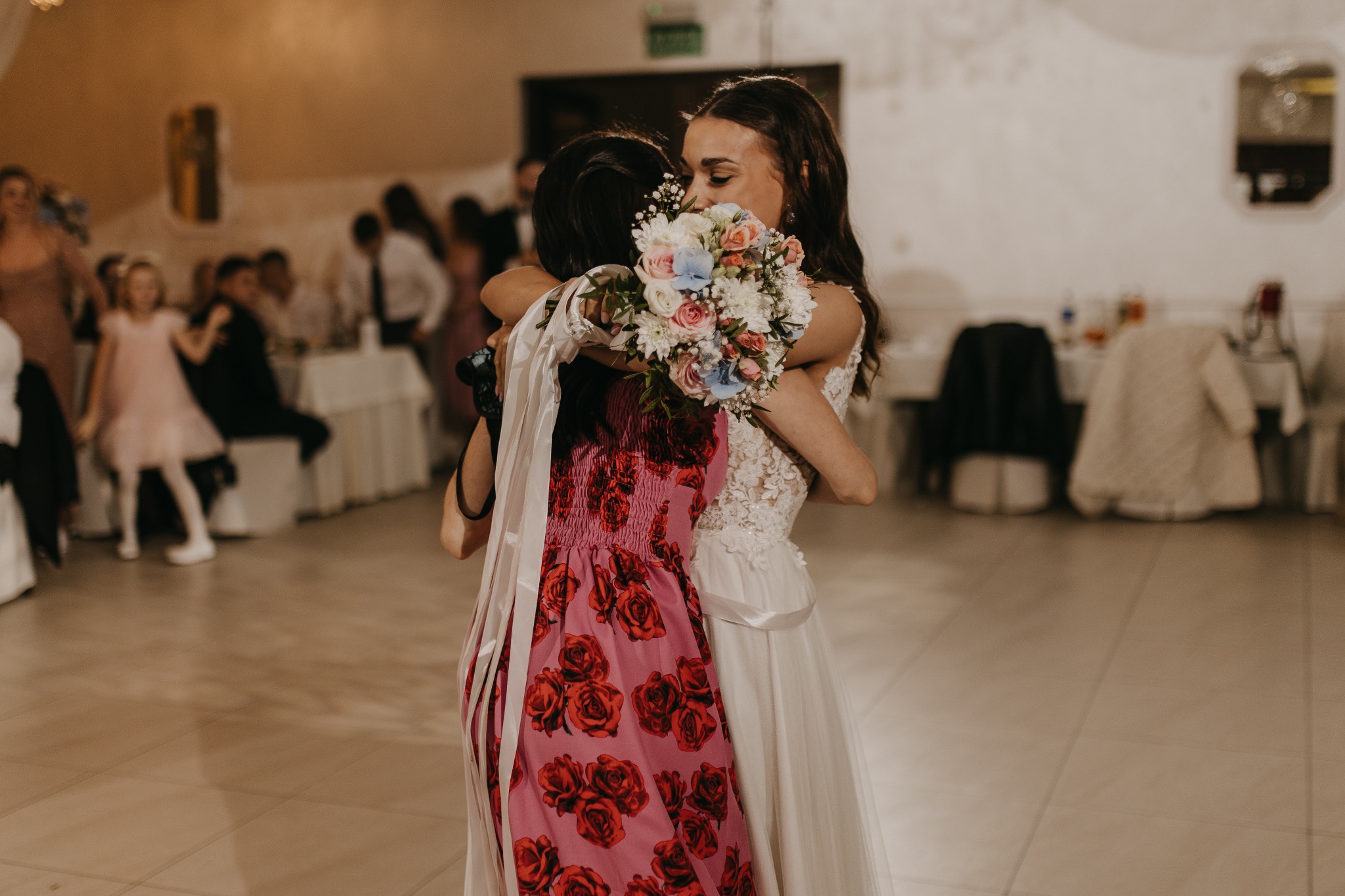 Panna młoda w białej sukni ślubnej z koronką i bukietem kwiatów obejmuje kobietę w różowej sukience w czerwone róże podczas wesela w sali bankietowej z gośćmi w tle.