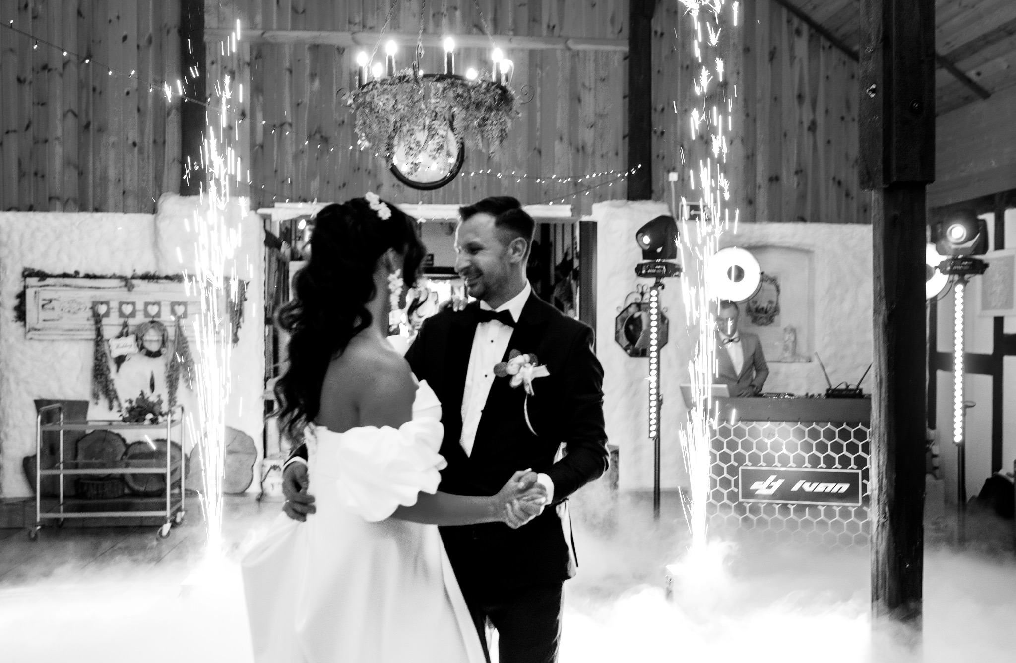 Para młoda w tańcu otoczona fontannami iskier i dymem, DJ w tle, rustykalne wnętrze z drewnianymi ścianami i dekoracyjnym oświetleniem.