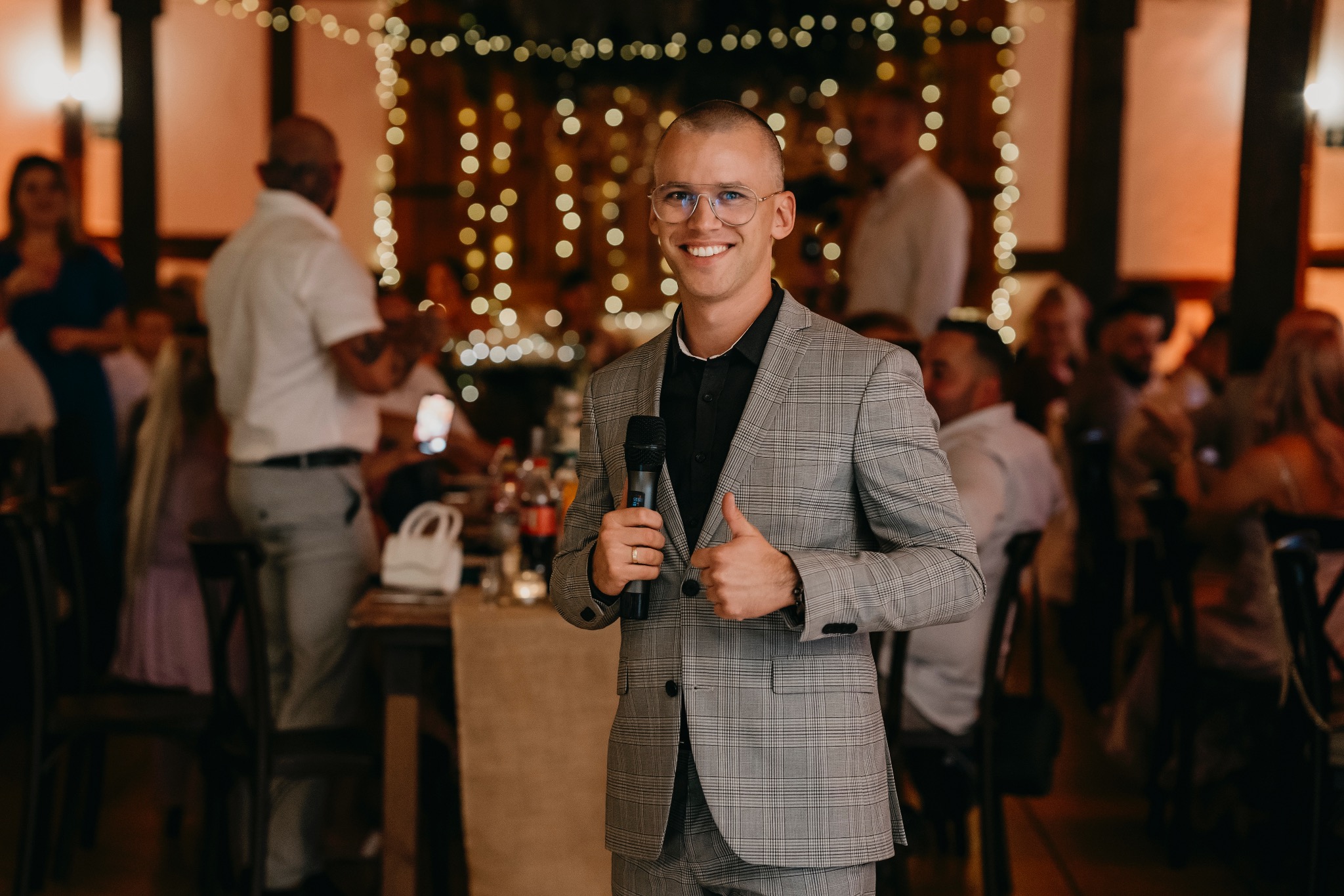 Uśmiechnięty mężczyzna w okularach i szarej kraciastej marynarce trzyma mikrofon, pokazując kciuk w górę na tle sali udekorowanej lampkami.