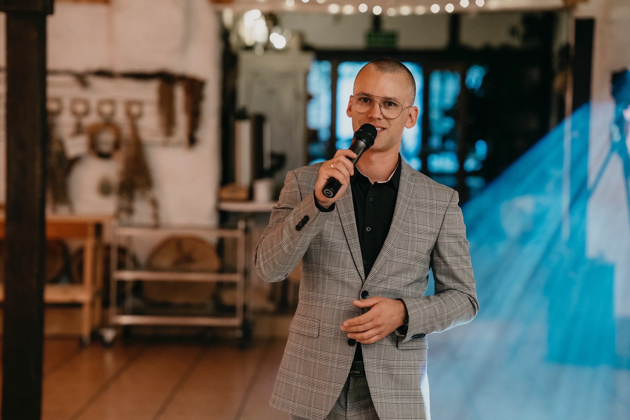 Mężczyzna w okularach i szarej kraciastej marynarce trzyma mikrofon, przemawiając na tle rozmytego wnętrza z dekoracjami i oświetleniem.