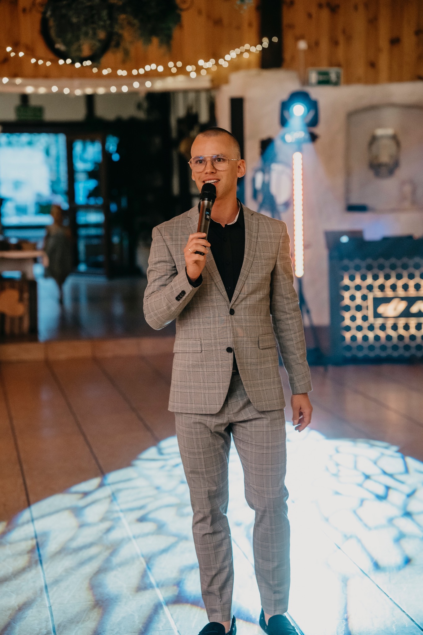 Mężczyzna w okularach i szarym garniturze w kratę trzyma mikrofon, przemawiając na tle sceny z oświetleniem LED.