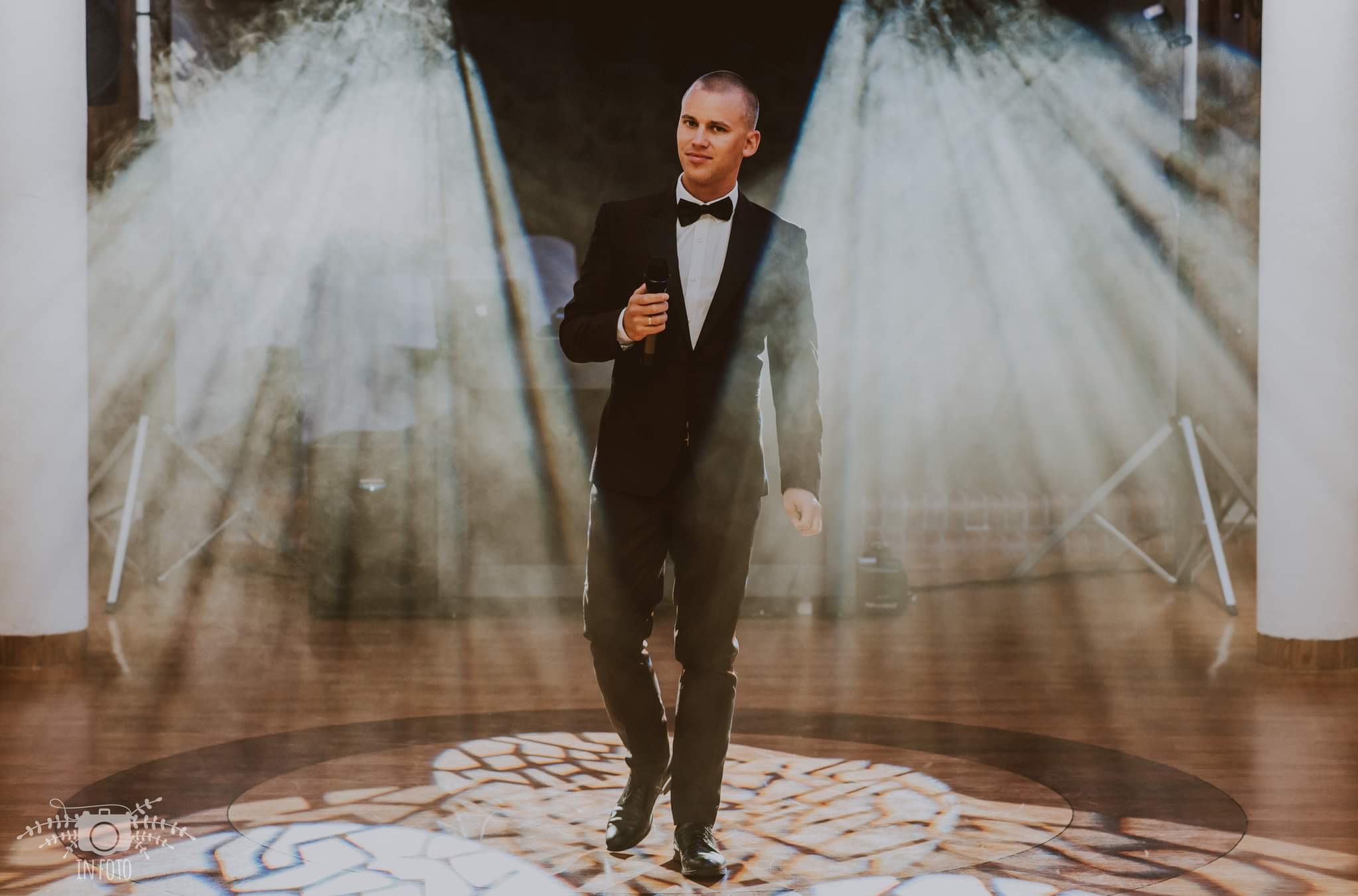 Elegancko ubrany mężczyzna w muszce, trzymający mikrofon, idzie po scenie oświetlonej reflektorami.