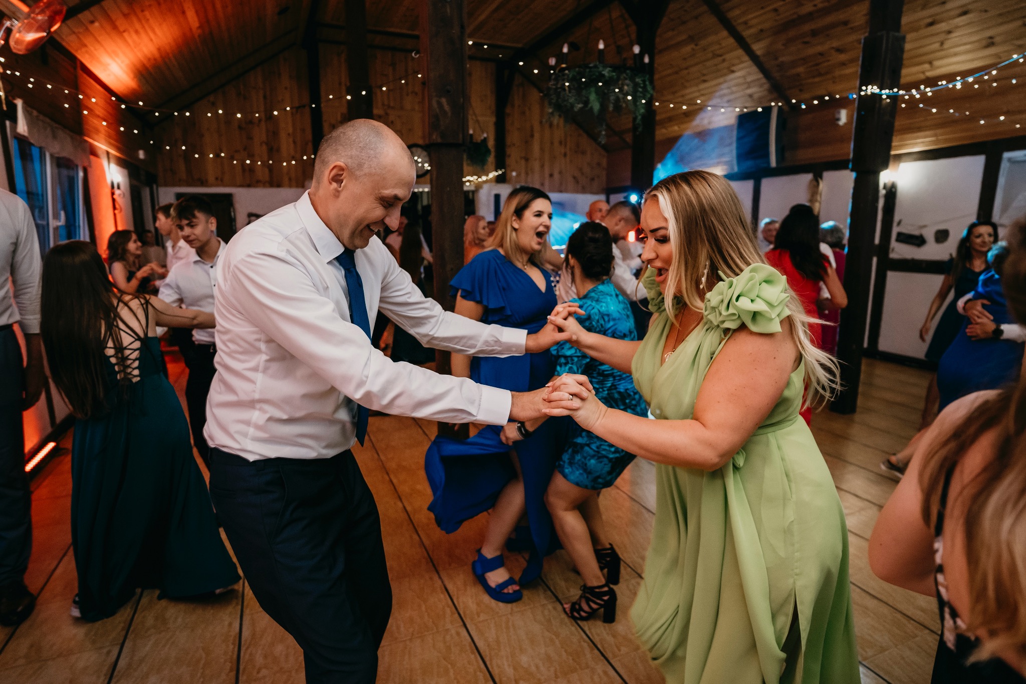 Goście weselni w trakcie tańca w sali z drewnianym sufitem i dekoracyjnymi lampkami.