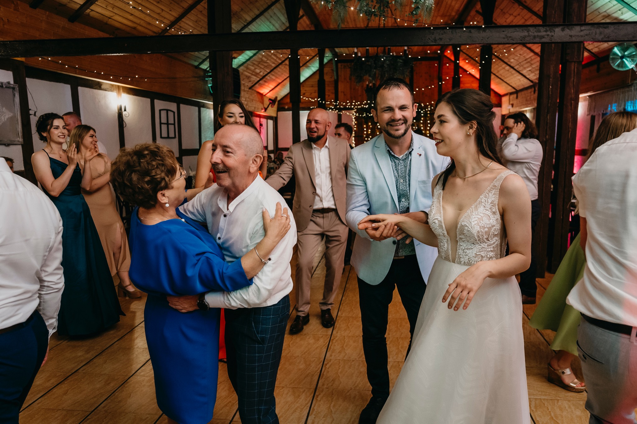 Weselna zabawa: panna młoda w koronkowej sukni i jasnym makijażu tańczy z gośćmi na parkiecie, drewniany sufit z dekoracyjnym oświetleniem w tle.