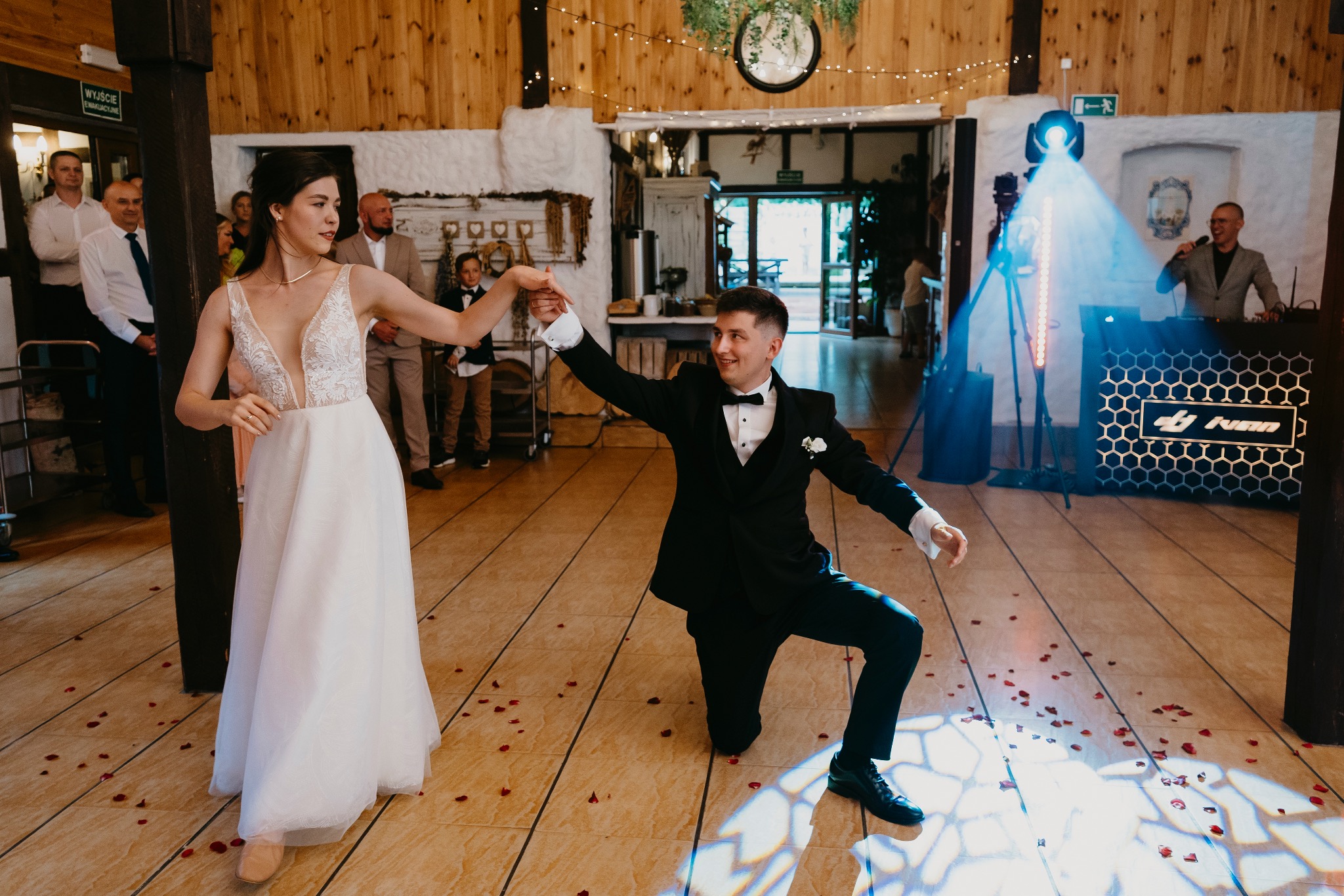Para młoda tańczy podczas wesela w Koszalinie, oświetlenie sceniczne, parkiet posypany płatkami róż, w tle wodzirej z mikrofonem.