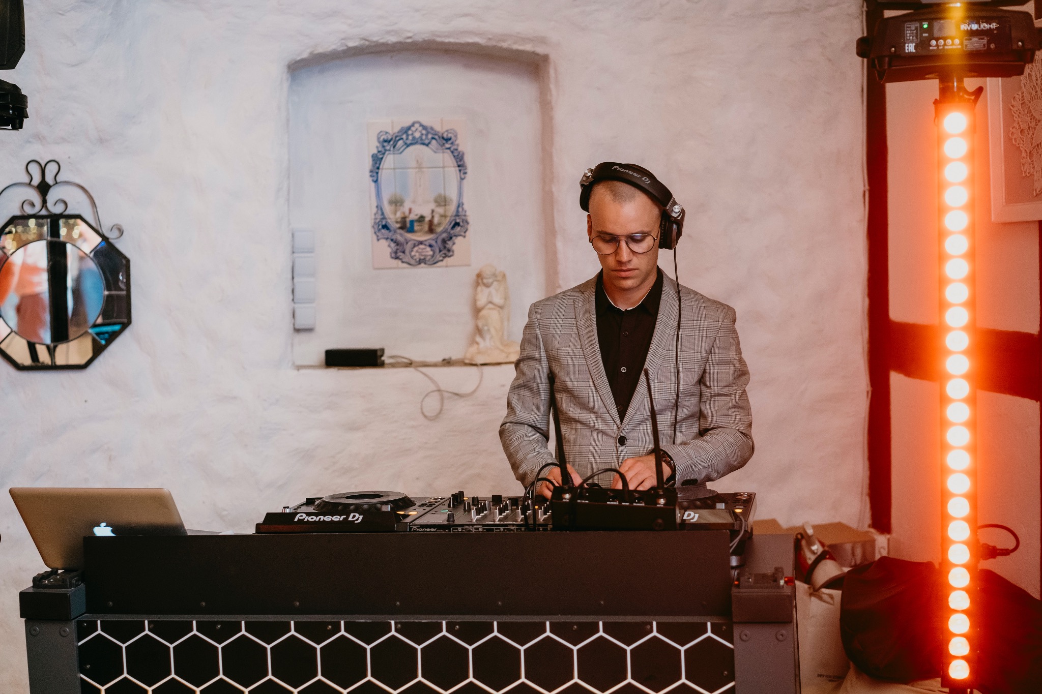 DJ w szarej marynarce w kratę, skupiony na mikserze Pioneer DJ, słuchawki na uszach, pomarańczowe światło w tle, rustykalne wnętrze z białymi ścianami i dekoracjami.