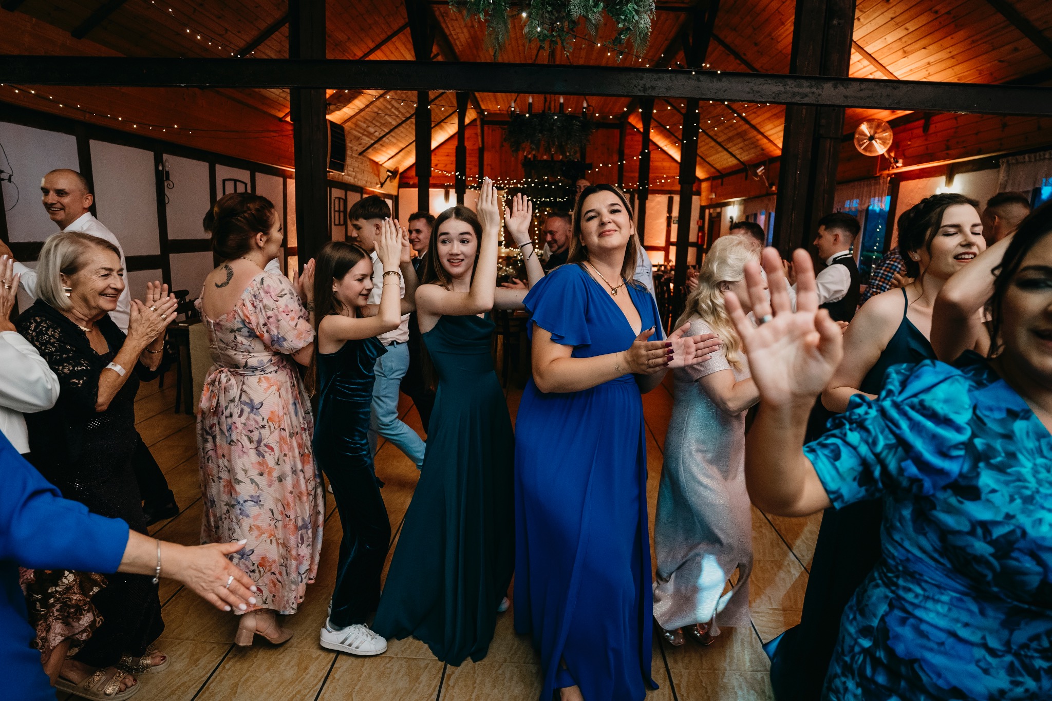 Grupa osób w eleganckich strojach tańczy i klaszcze w dłonie w rustykalnej sali z drewnianym sufitem i oświetleniem girlandami, podczas imprezy.