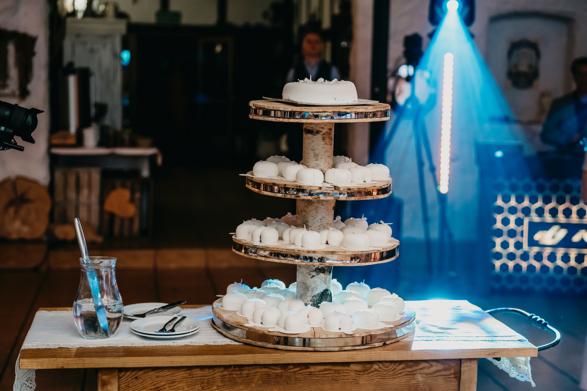 Czteropoziomowy tort weselny na drewnianym stojaku z brzozy, małe białe babeczki i duży tort na górze, nóż i sztućce na stole, niebieskie oświetlenie sceniczne w tle.