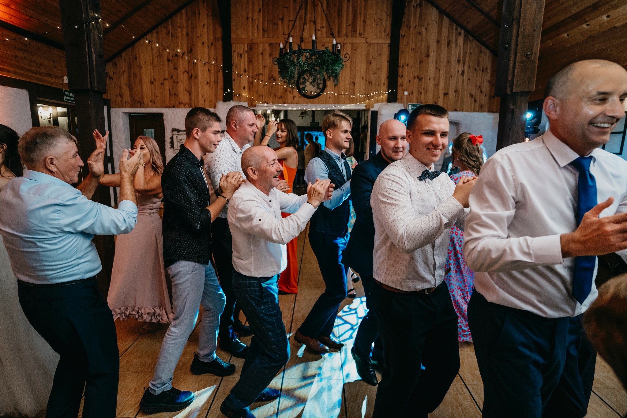 Uczestnicy wesela tańczą i klaszczą w rytm muzyki w rustykalnej sali z drewnianymi ścianami i dekoracyjnym żyrandolem.