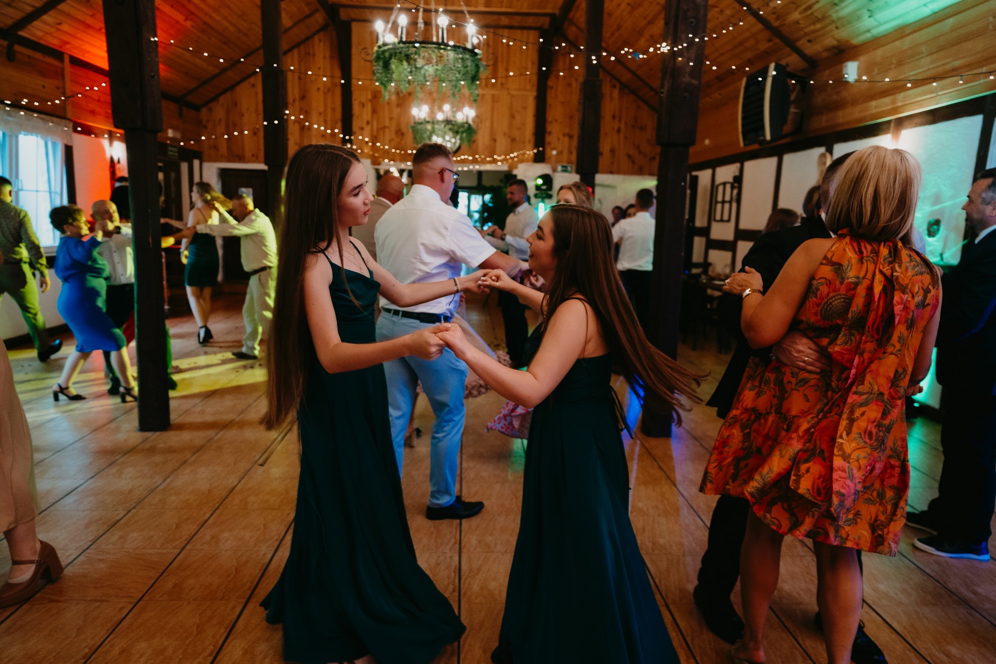 Goście weselni tańczą na parkiecie w drewnianej sali z dekoracyjnym żyrandolem i lampkami.