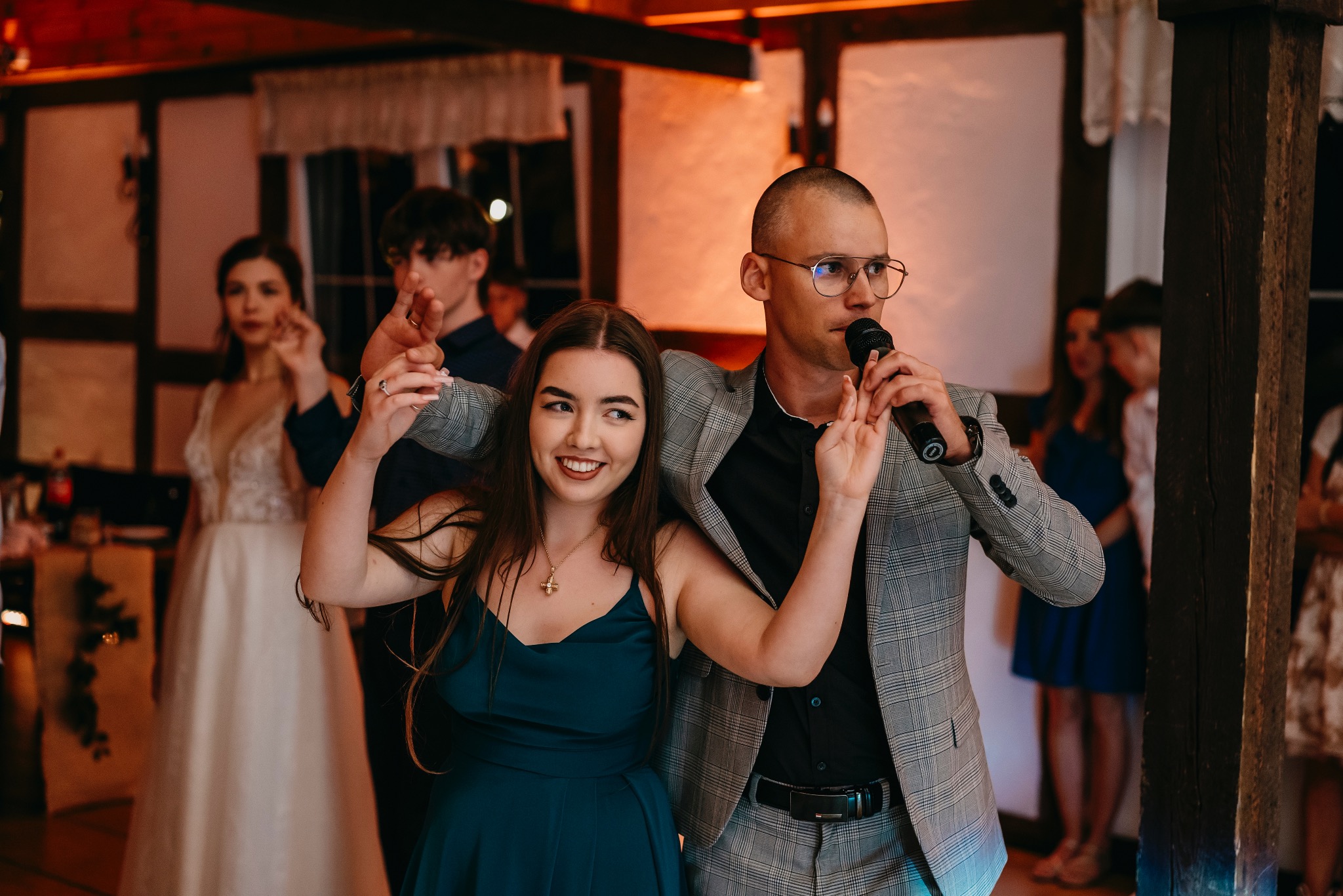 Wokalista w okularach i kraciastej marynarce śpiewa do mikrofonu, obejmując uśmiechniętą kobietę w szmaragdowej sukience podczas występu na tle gości weselnych.