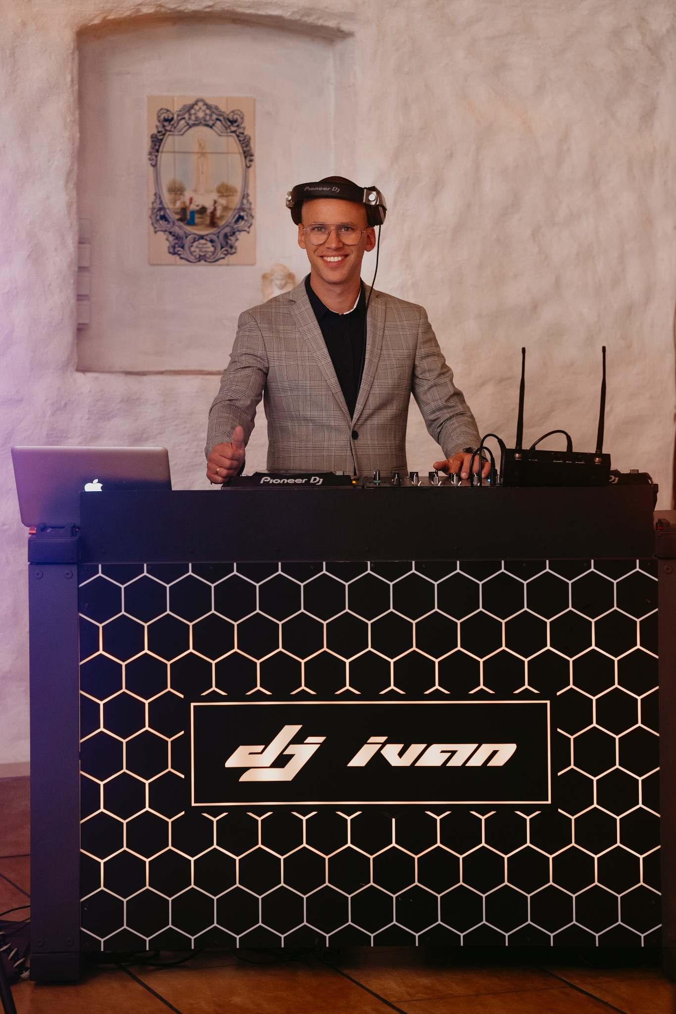 Uśmiechnięty DJ w okularach i marynarce, z słuchawkami na głowie, stoi za konsoletą z laptopem, dając kciuk w górę, w tle biała ściana z dekoracyjną płytką.