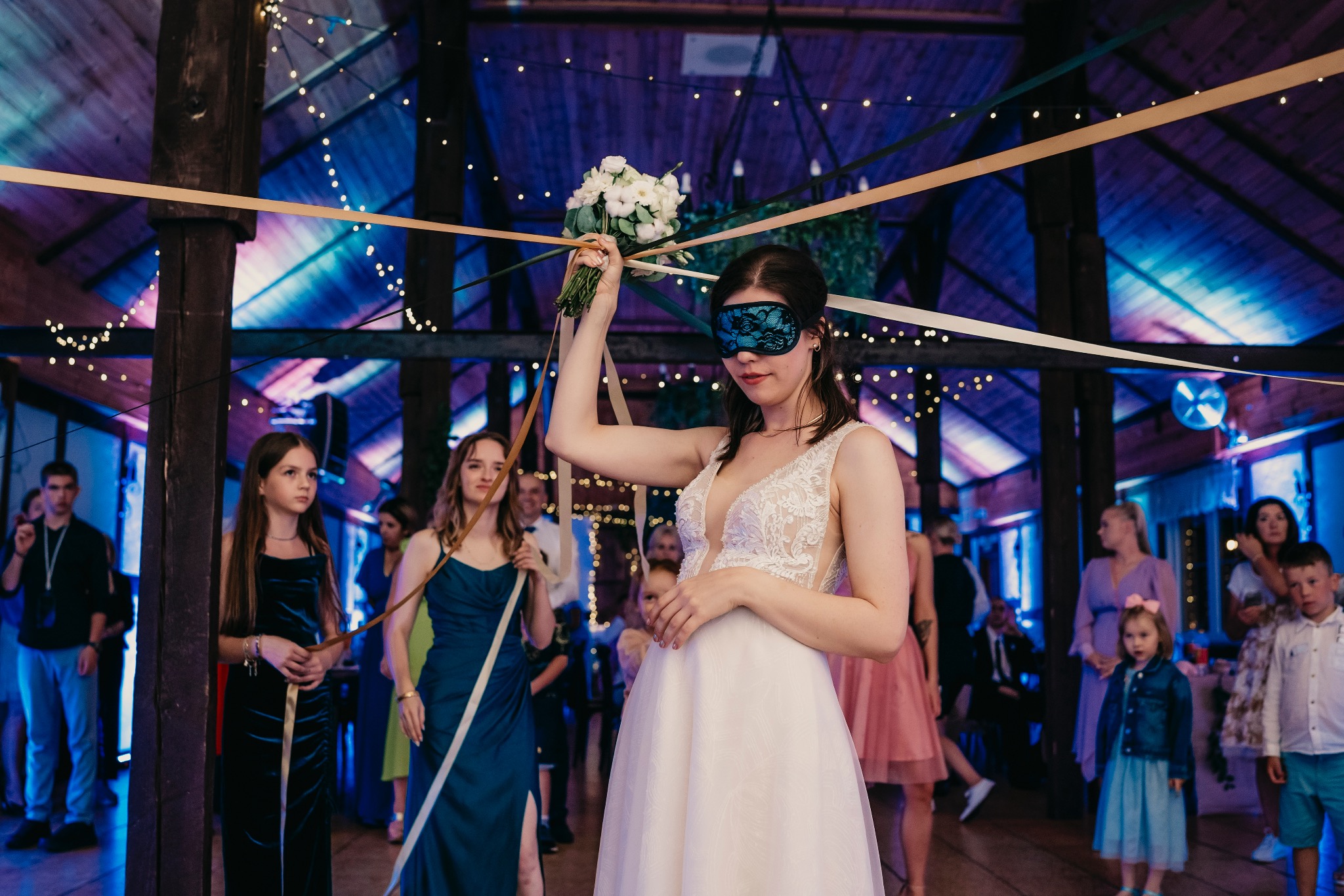 Panna młoda z zasłoniętymi oczami trzyma bukiet podczas tradycyjnego weselnego oczepin w drewnianej sali z dekoracjami świetlnymi i wstążkami.