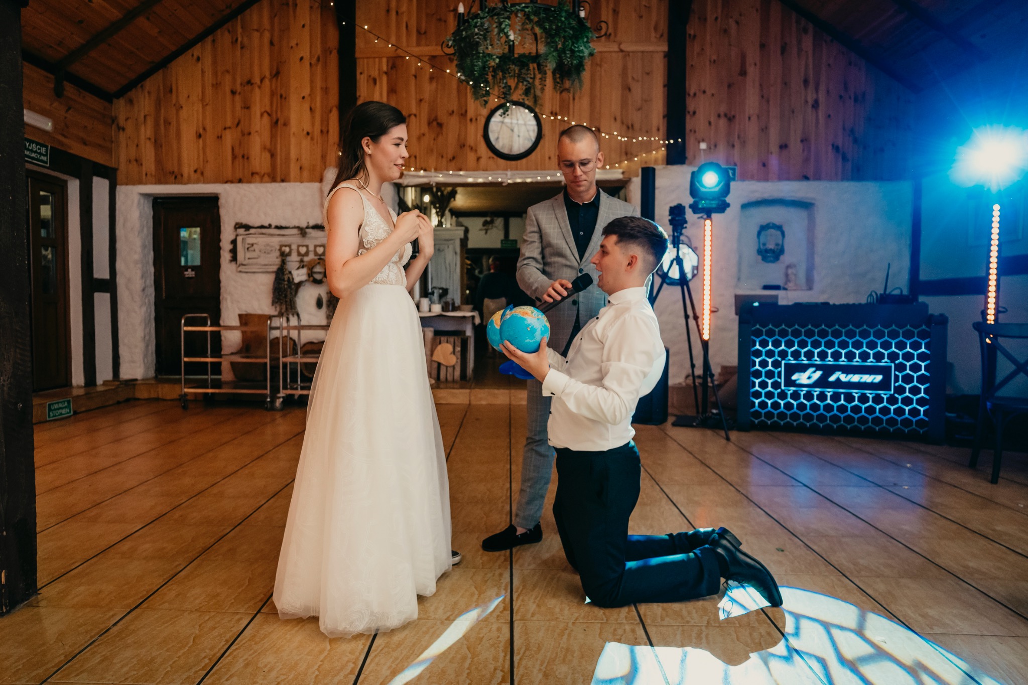 Mężczyzna klęczy z globusem przed panną młodą podczas uroczystości, konferansjer z mikrofonem w tle, dekoracje świetlne i drewniane wykończenie sali.