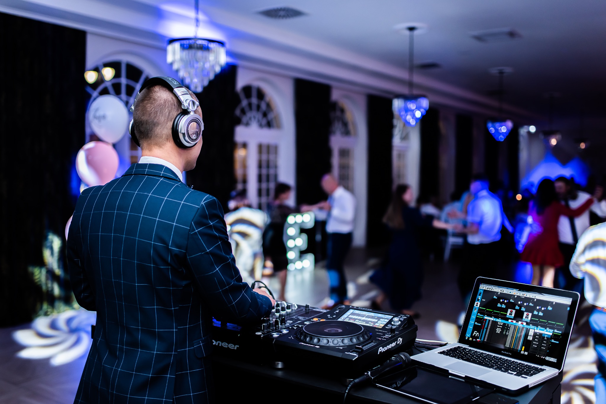 DJ w granatowym garniturze w kratę, tyłem, miksuje muzykę na profesjonalnym sprzęcie Pioneer DJ podczas imprezy tanecznej; w tle rozświetlona sala balowa z tańczącymi ludźmi i dekoracjami.