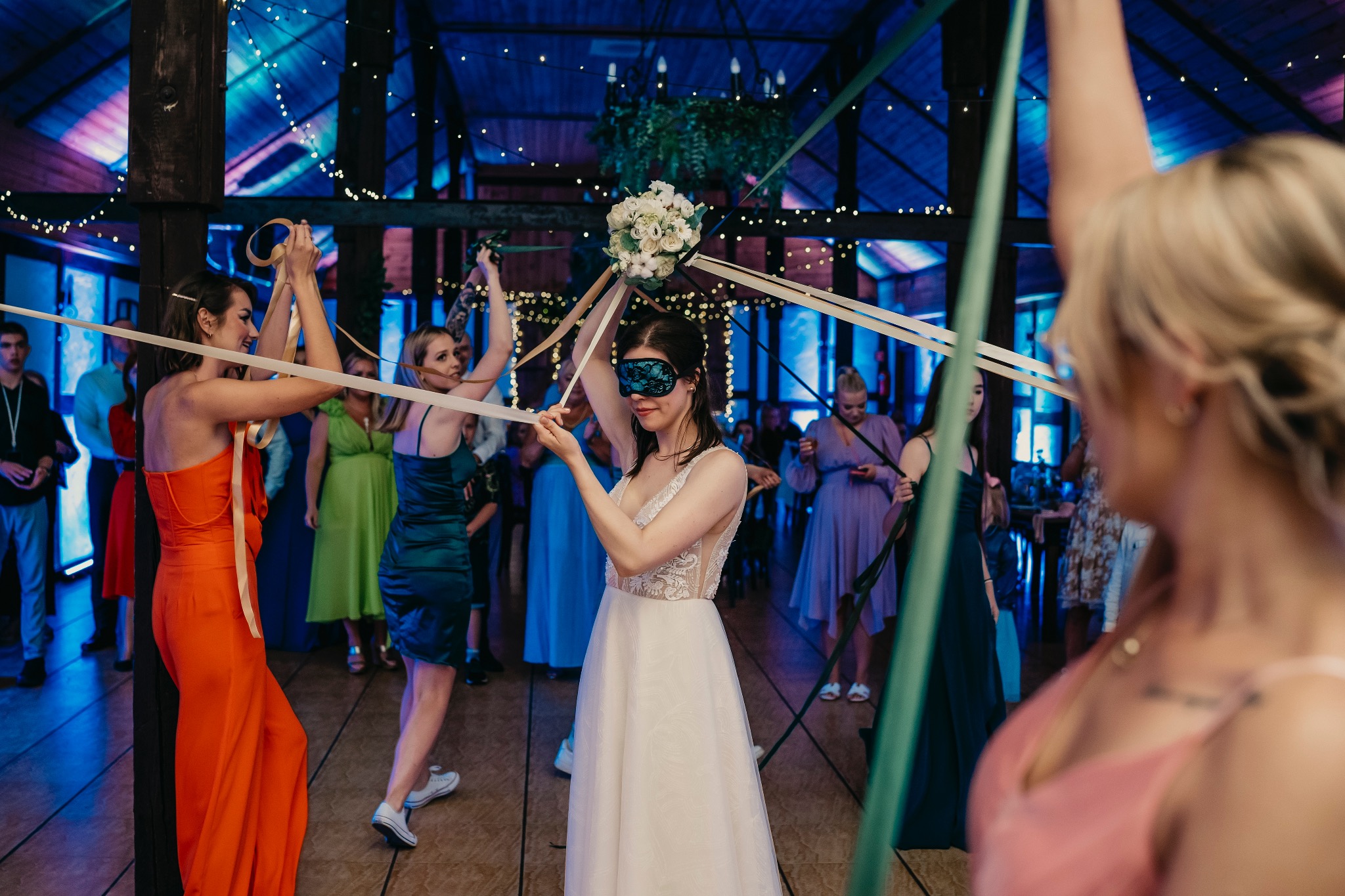 Panna młoda z zasłoniętymi oczami, trzymająca bukiet, uczestniczy w tradycyjnej zabawie weselnej z wstążkami, otoczona gośćmi w sali z drewnianą konstrukcją i dekoracyjnym oświetleniem.
