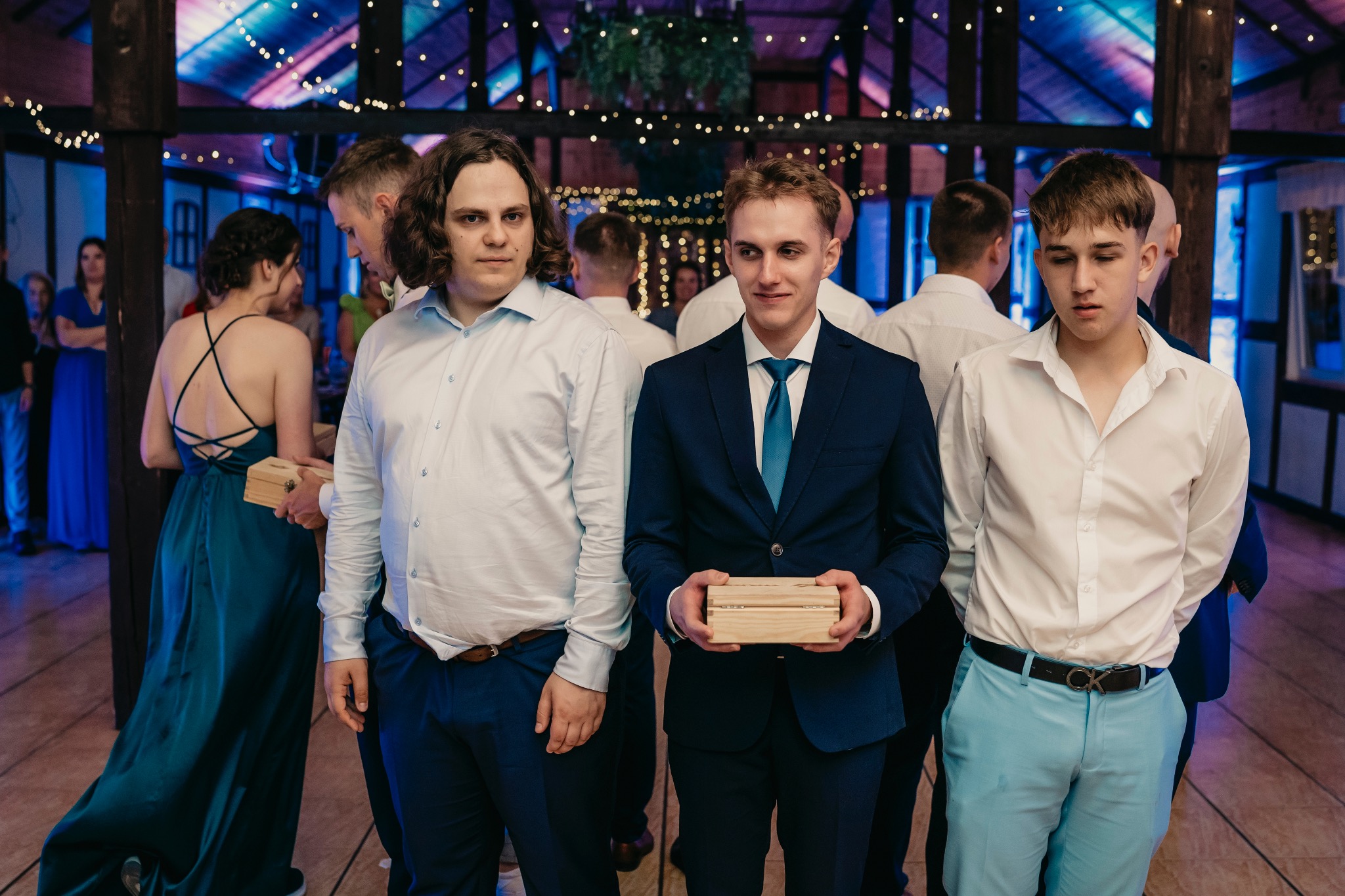Trzech mężczyzn w różnym wieku i ubraniach trzyma małe drewniane pudełka na tle dekoracji sali weselnej z oświetleniem.