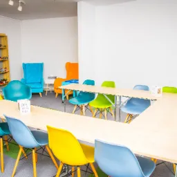 Jasna sala konferencyjna z drewnianym stołem w kształcie litery U otoczonym kolorowymi krzesłami, w tle regał z zabawkami i dwa fotele w odcieniach niebieskiego i pomarańczowego.