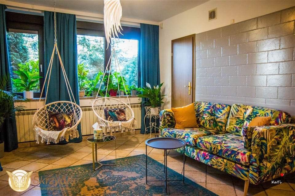 Jasne wnętrze pokoju z dwoma wiszącymi, plecionymi fotelami, wzorzystą sofą, zielonym dywanem i roślinami w oknach, oświetlone nowoczesną lampą.