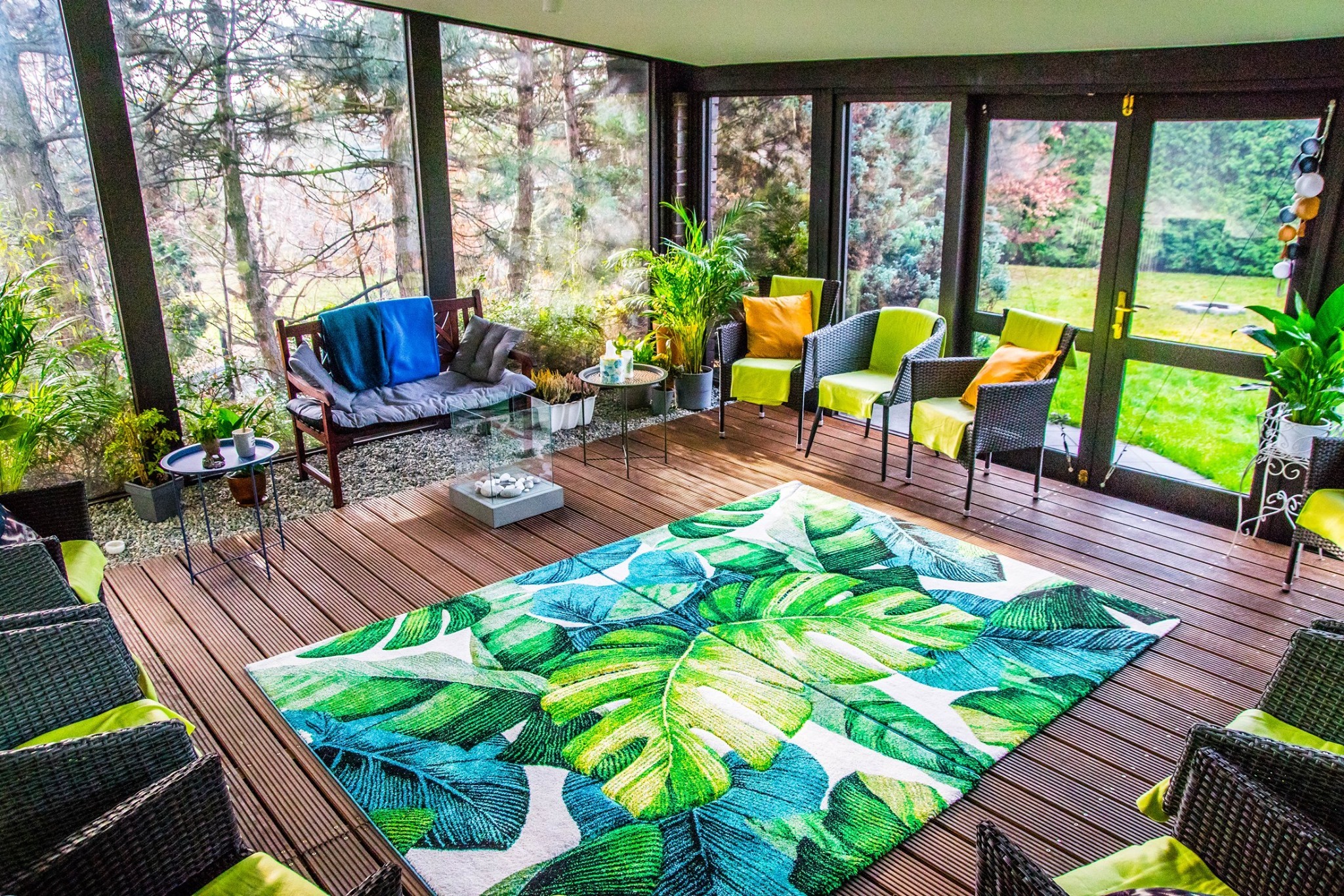 Zadaszony taras z drewnianą podłogą, meblami rattanowymi z kolorowymi poduszkami, dywanem w motywie liści i widokiem na ogród przez przeszklone ściany.