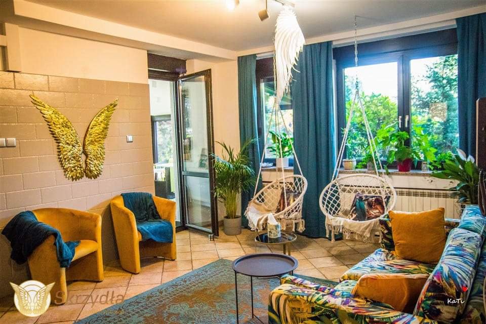 Jasne wnętrze pokoju z dwoma wiszącymi fotelami hamakowymi, dwoma żółtymi fotelami, kanapą w tropikalne wzory, dywanem i złotymi skrzydłami dekoracyjnymi na ścianie.