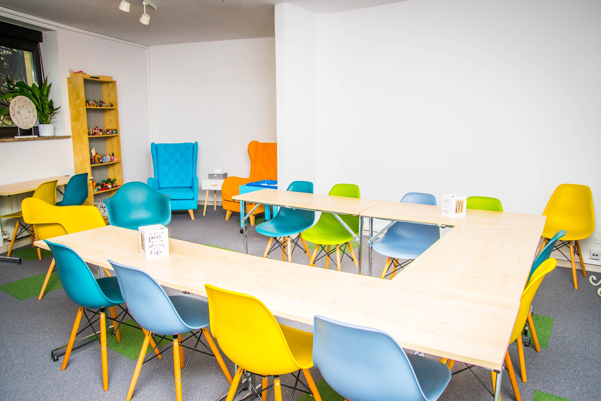 Jasna sala konferencyjna z drewnianym stołem w kształcie litery U otoczonym kolorowymi krzesłami, w tle regał z zabawkami i dwa fotele w odcieniach niebieskiego i pomarańczowego.