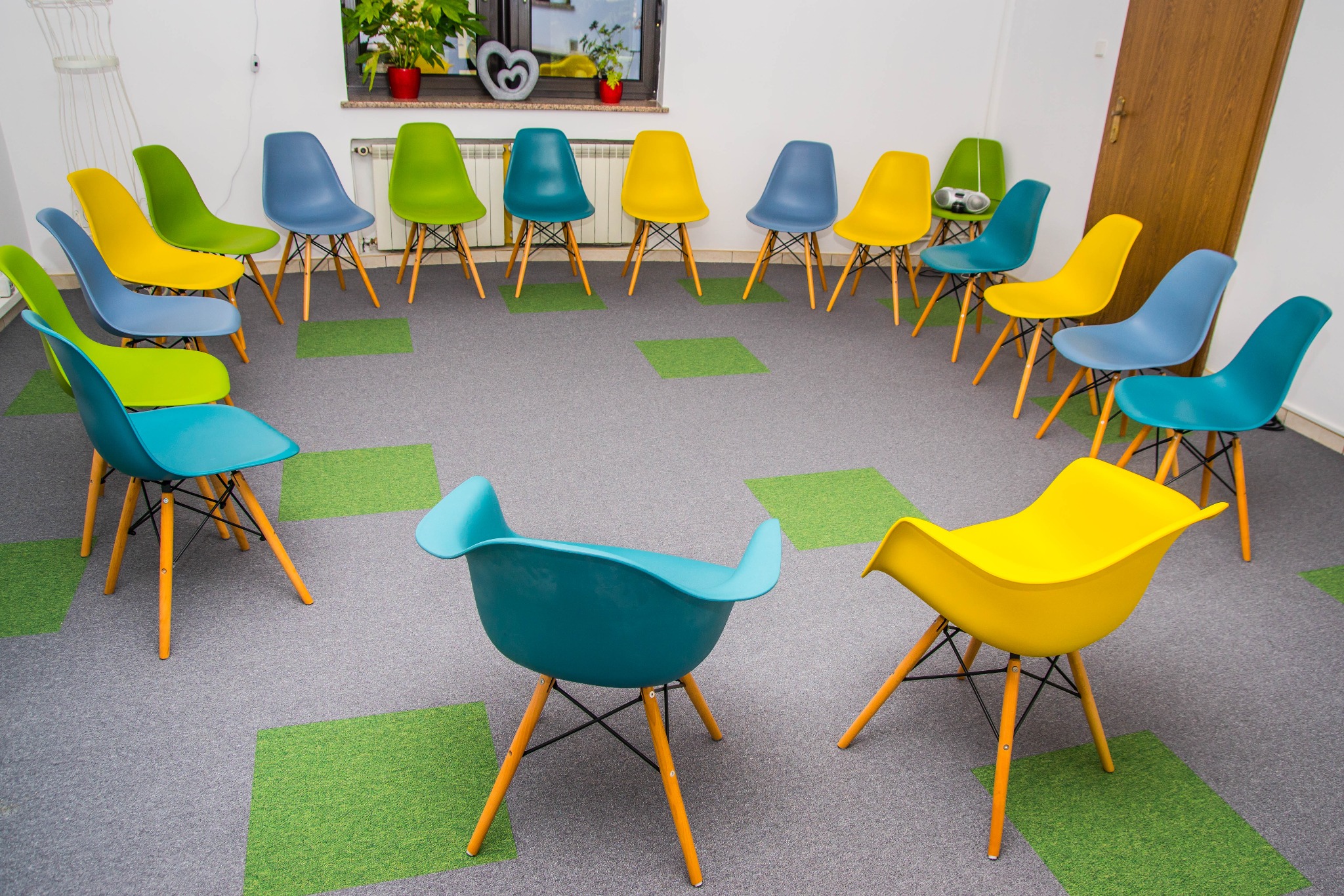 Sala z krzesłami ustawionymi w okrąg, przygotowana do warsztatów lub sesji terapeutycznej, z kolorowymi krzesłami i wykładziną w szaro-zielone kwadraty.