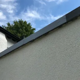 Fragment elewacji budynku z szarą listwą okapową i świeżo pomalowanym fragmentem, na tle błękitnego nieba z chmurami i zielonego drzewa.