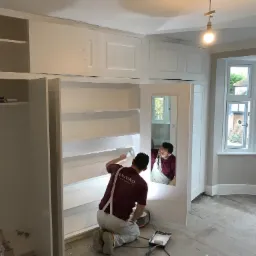 Malarz w bordowej koszulce z logo, klęczący maluje na biało wnętrze szafy wnękowej, widoczne narzędzia malarskie i odbicie malarza w lustrze.