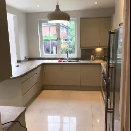 Jasna, nowoczesna kuchnia z beżowymi szafkami, szarym blatem i płytkami podłogowymi w odcieniach kremu, widok na ogród przez okno nad zlewem.