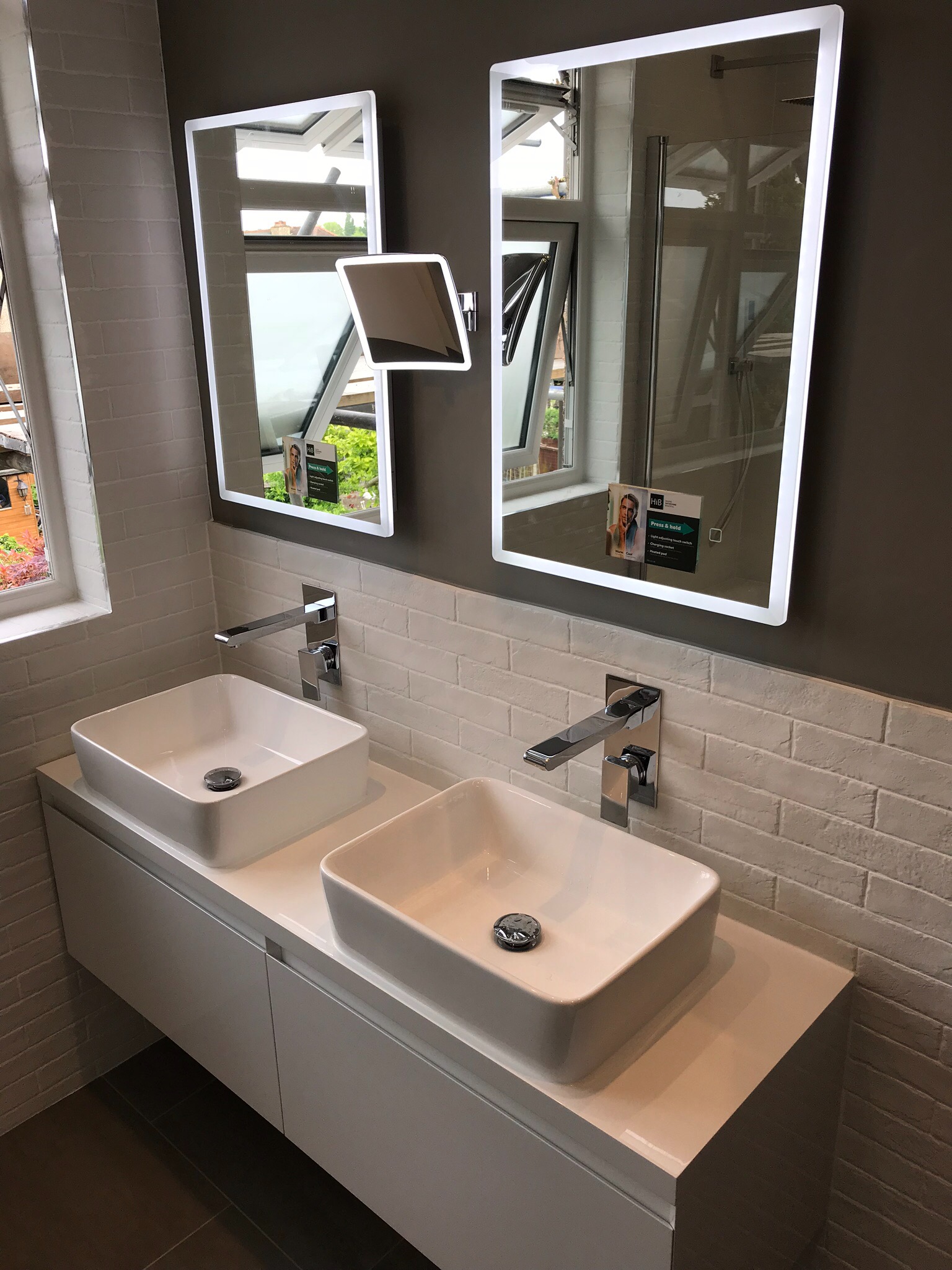 Nowoczesna łazienka z dwoma kwadratowymi umywalkami nablatowymi, chromowanymi bateriami i podświetlanymi lustrami LED, wykończona białą cegiełką i szarą ścianą.
