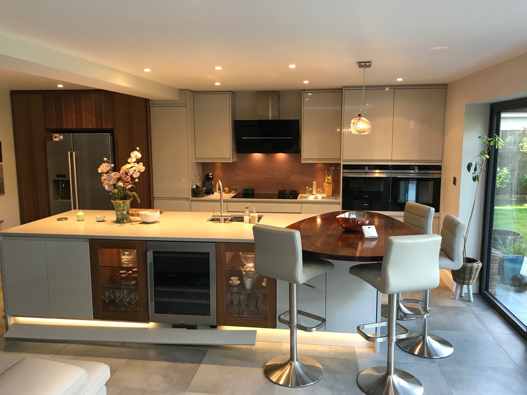 Nowoczesna kuchnia z wyspą, drewnianym blatem barowym, szarymi szafkami, stalową lodówką i oświetleniem LED pod szafkami. Widoczne trzy krzesła barowe i przeszklona witryna z kieliszkami.