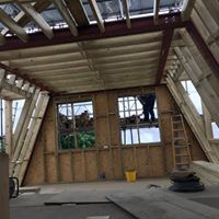 Wnętrze poddasza w trakcie prac wykończeniowych, widoczne drewniane elementy konstrukcyjne ścian i dachu, dwa okna z widokiem na zieleń, pracownik na drabinie, żółte wiadro i płyty OSB na podłodze.