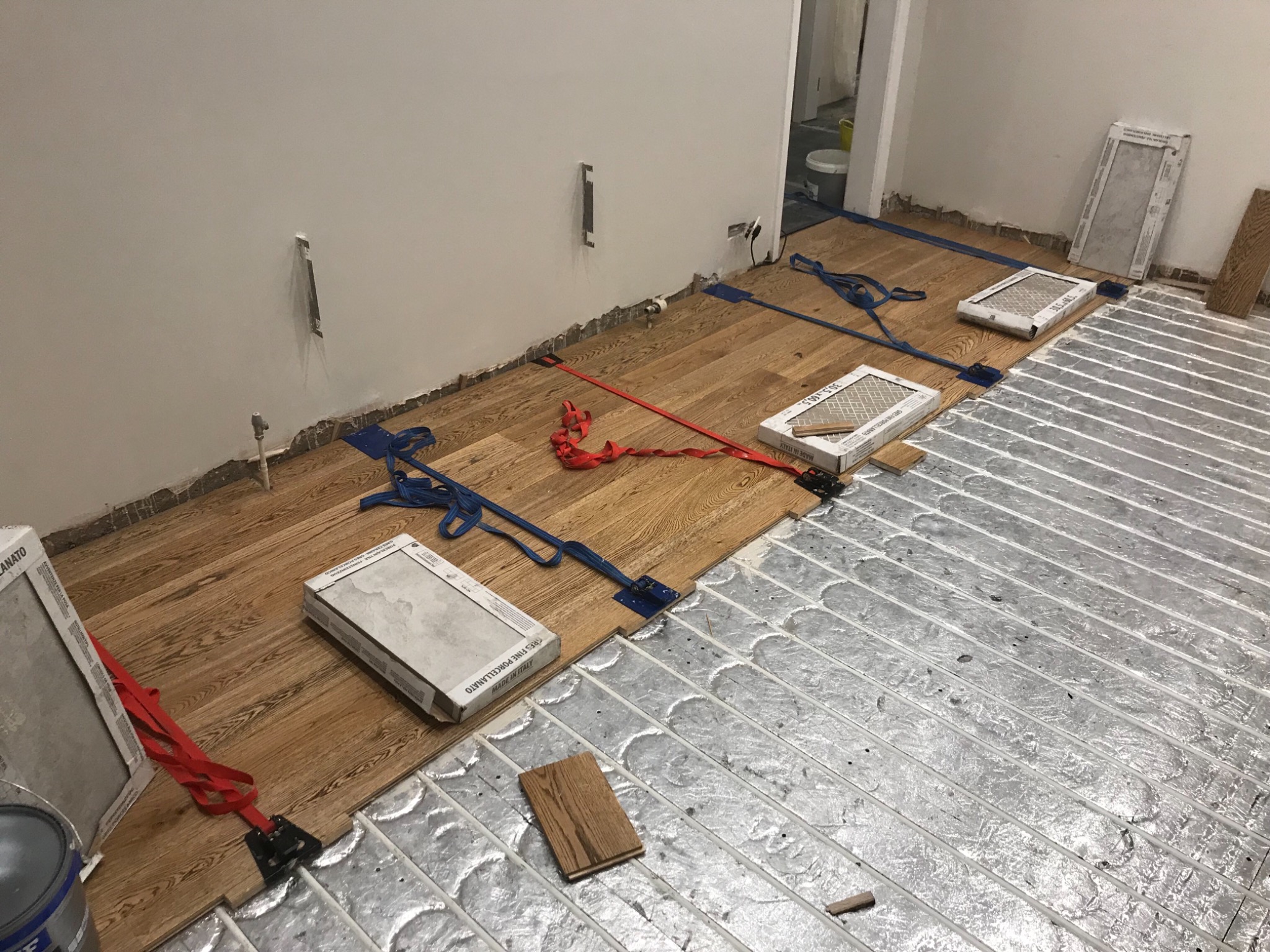 Montaż drewnianej podłogi na ogrzewaniu podłogowym, widoczne listwy przypodłogowe, paski mocujące i pudełka z płytkami.
