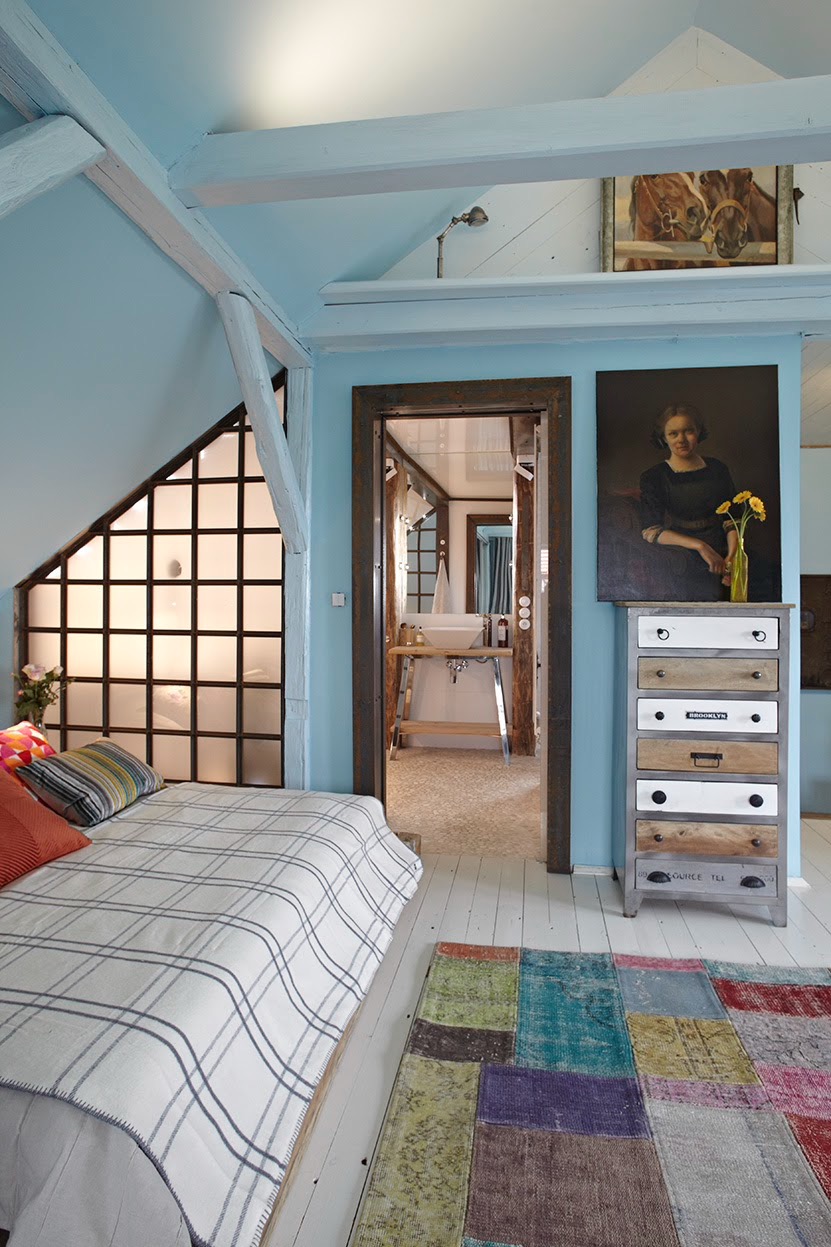 Jasny pokój na poddaszu z łóżkiem, patchworkowym dywanem i widokiem na łazienkę przez drzwi z surowego drewna; na ścianie obraz z kobietą i komoda z szufladami w stylu vintage.
