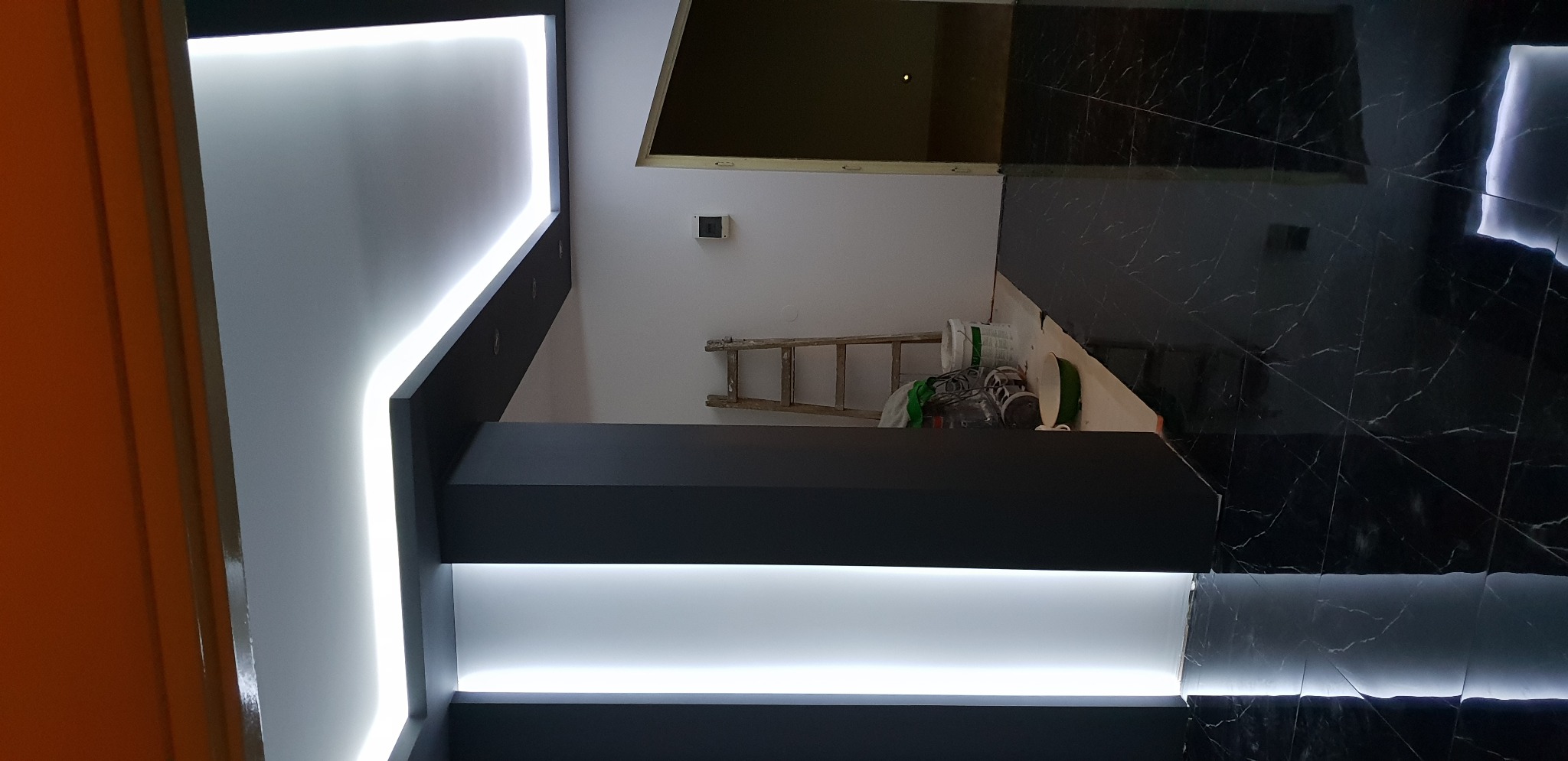 Częściowy widok wnętrza w trakcie prac: czarna zabudowa z podświetleniem LED, marmurowe płytki podłogowe, drabina oparta o ścianę, narzędzia i materiały budowlane w tle.