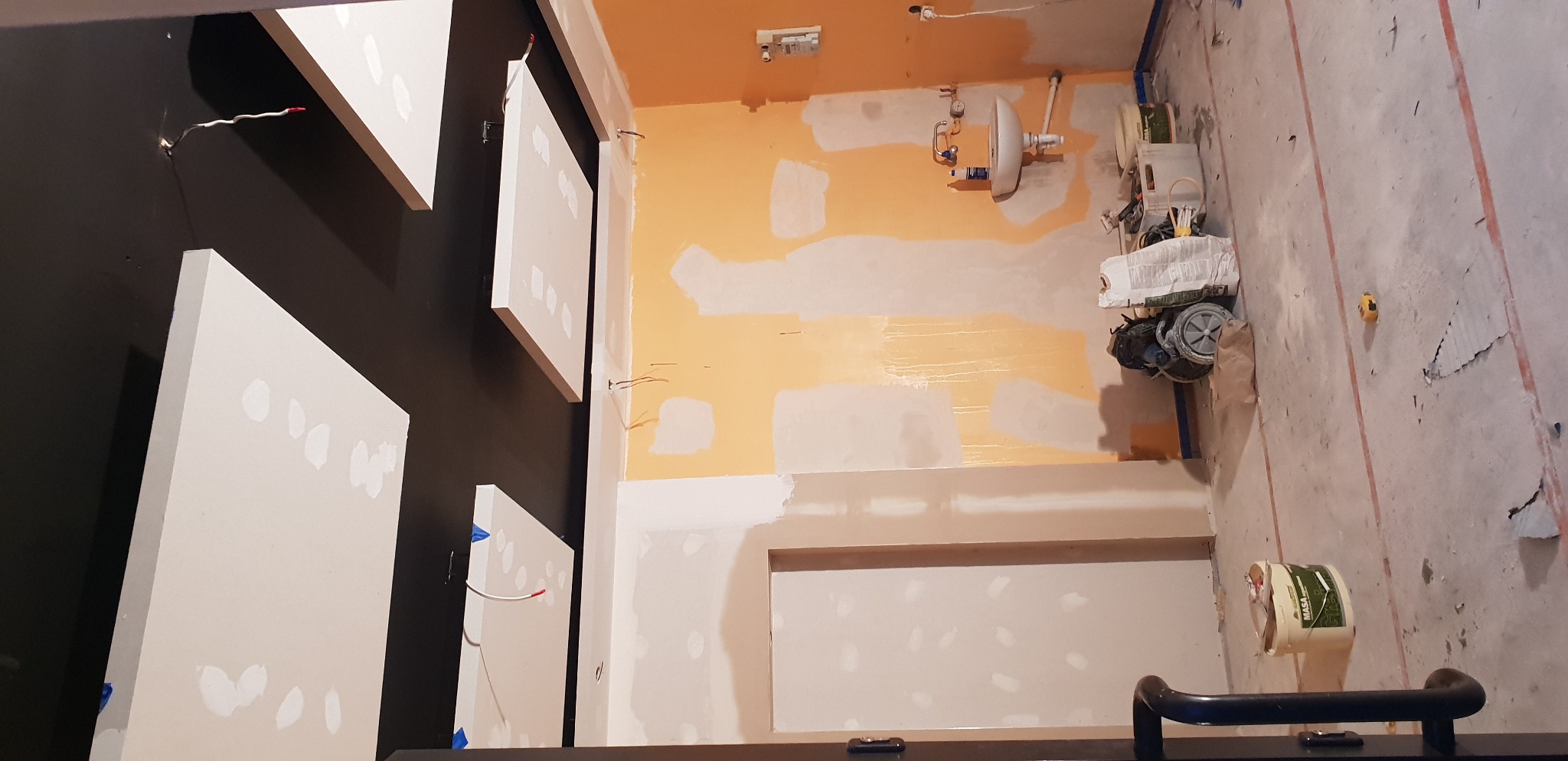 Wnętrze w trakcie prac wykończeniowych: ściany częściowo pokryte szpachlą i pomalowane na żółto, widoczne kable elektryczne, narzędzia budowlane i płyty gipsowo-kartonowe na czarnej ścianie.