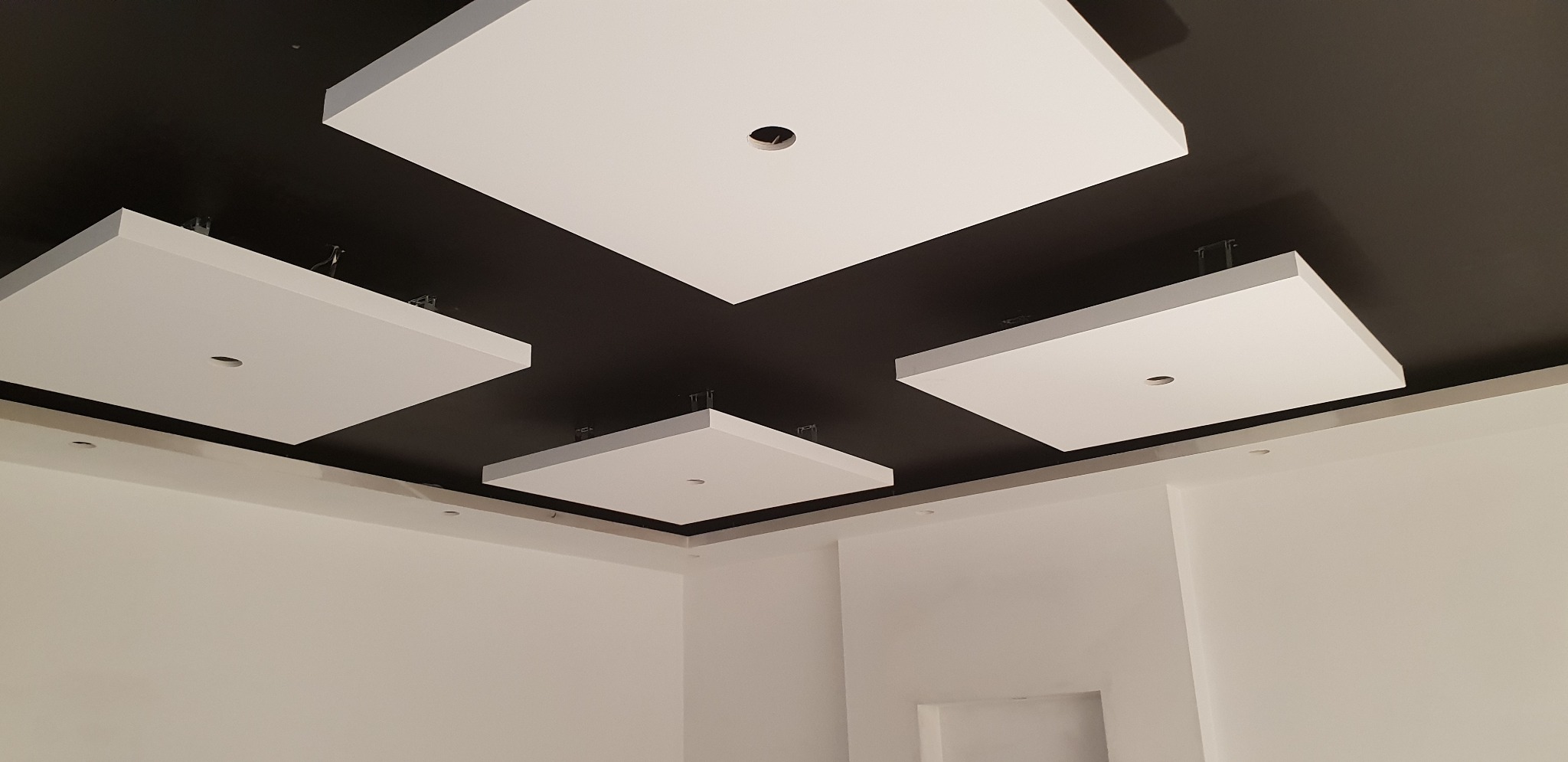 Nowoczesny sufit podwieszany z białymi kwadratowymi panelami na czarnym tle, widoczne metalowe mocowania, minimalistyczny design w jasnym pomieszczeniu.