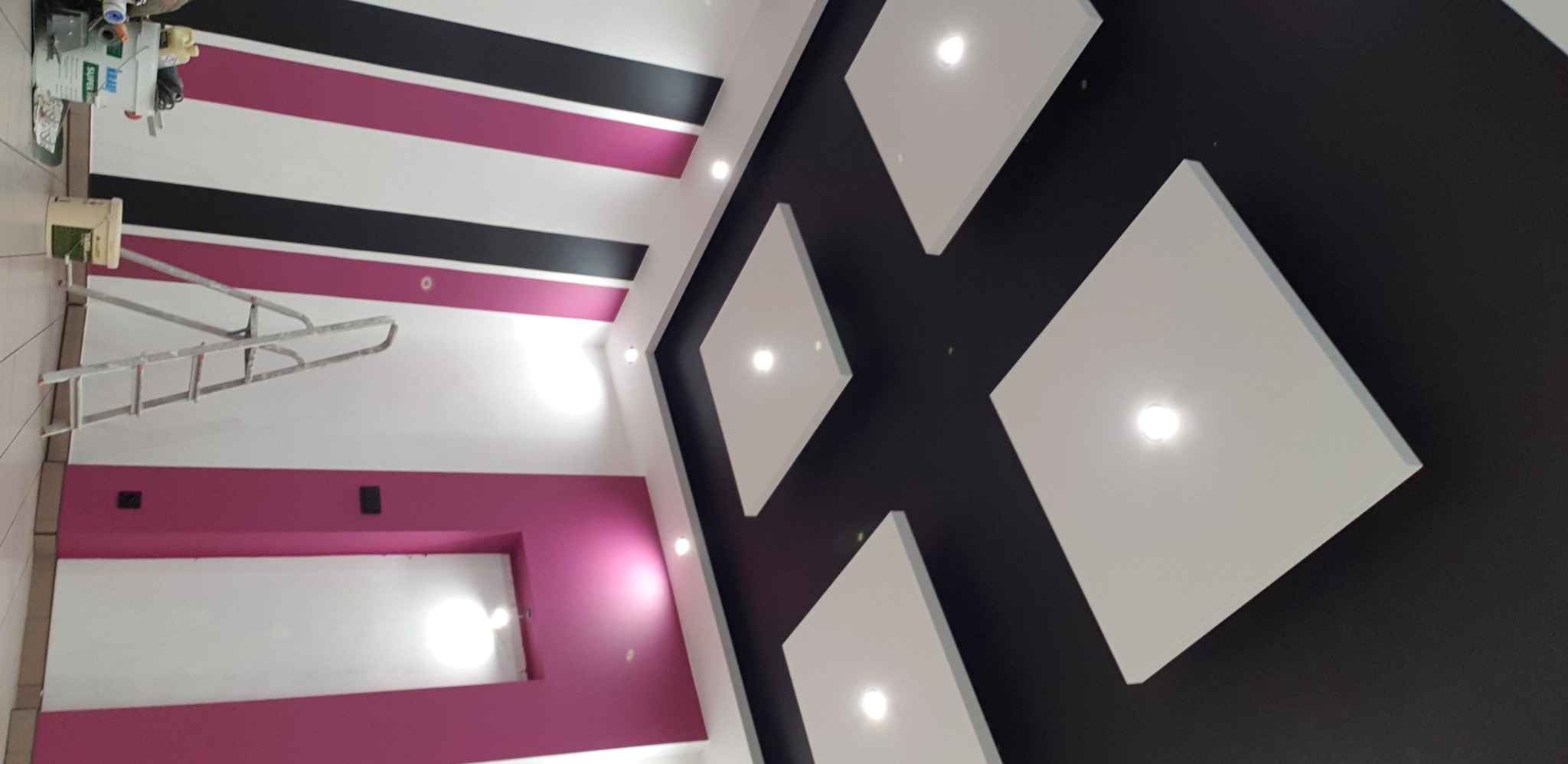 Wnętrze po odświeżeniu: ściany w biało-różowe pasy, czarny sufit z kwadratowymi panelami oświetleniowymi, drabina malarska i puszki z farbą w rogu.