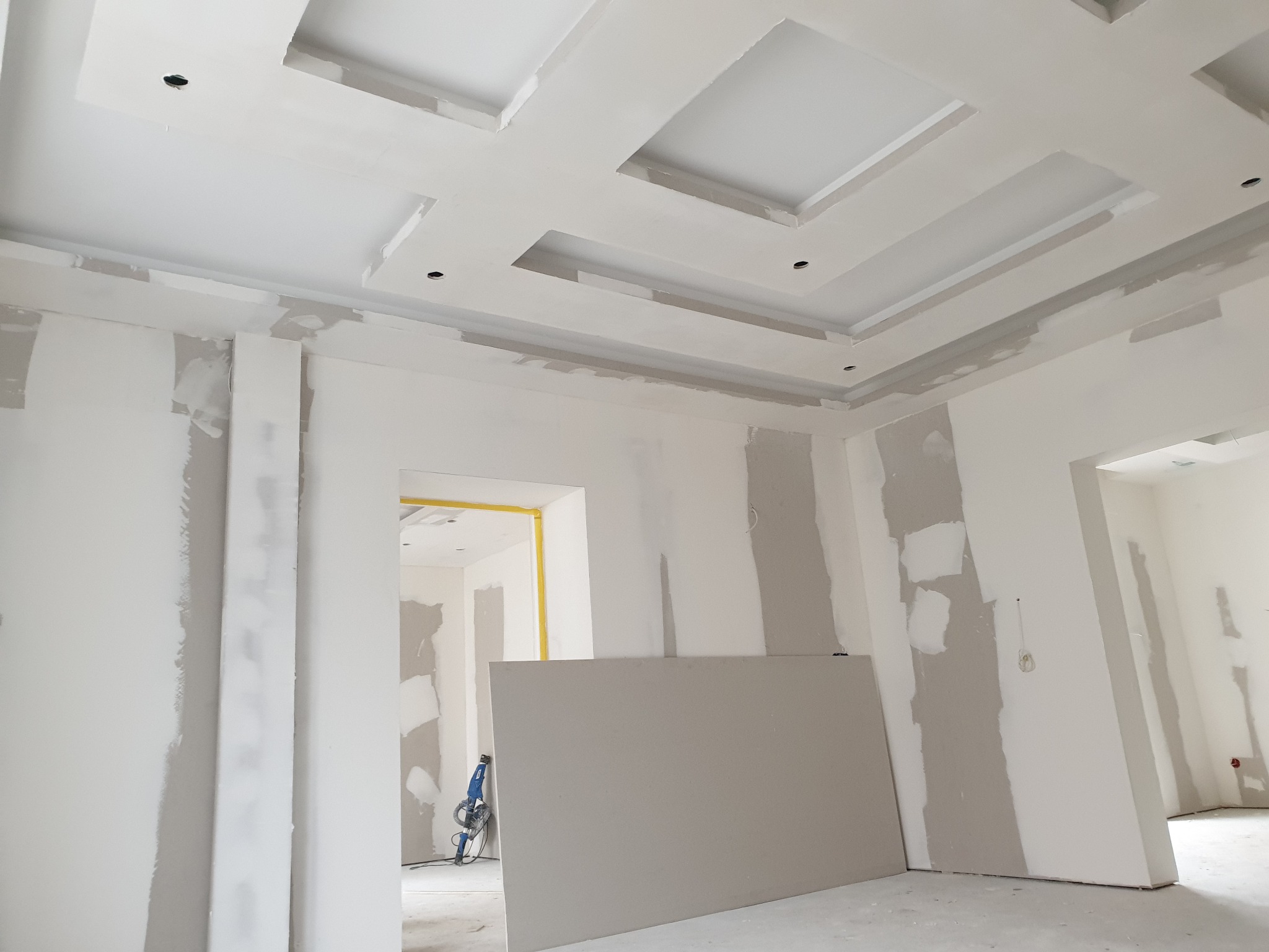 Wnętrze w trakcie remontu: widoczne ściany po szpachlowaniu, sufit z dekoracyjnymi wgłębieniami i punktami oświetleniowymi, płyta gipsowa oparta o ścianę i szlifierka do gipsu w tle.