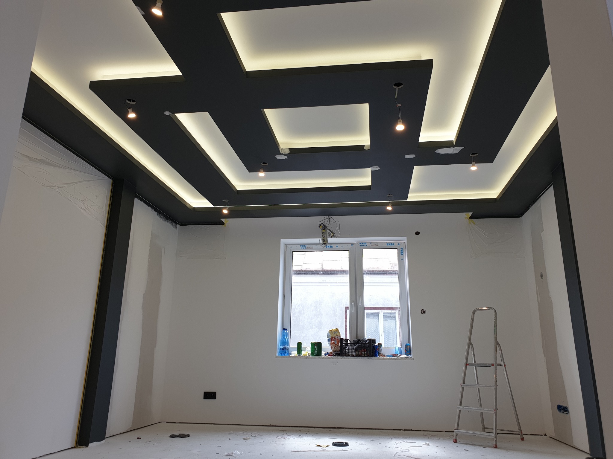 Wnętrze w trakcie prac wykończeniowych, widoczny sufit podwieszany z oświetleniem LED, ściany przygotowane do malowania, okno z materiałami budowlanymi na parapecie, drabina aluminiowa.