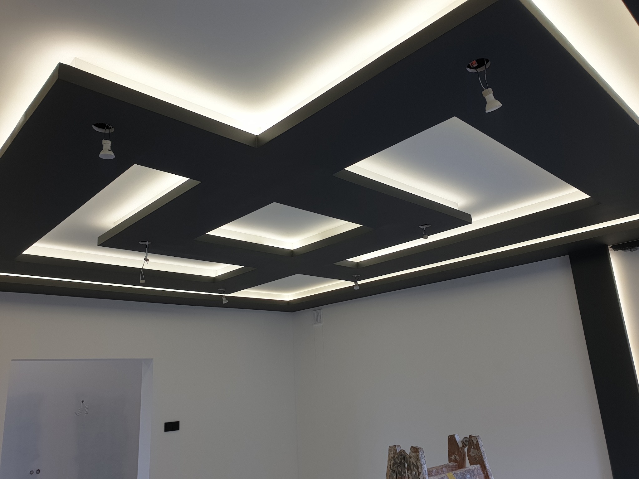 Nowoczesny sufit podwieszany z oświetleniem LED, pomalowany na ciemny kolor, z widocznymi punktami oświetleniowymi i fragmentem drabiny malarskiej w dolnej części kadru.