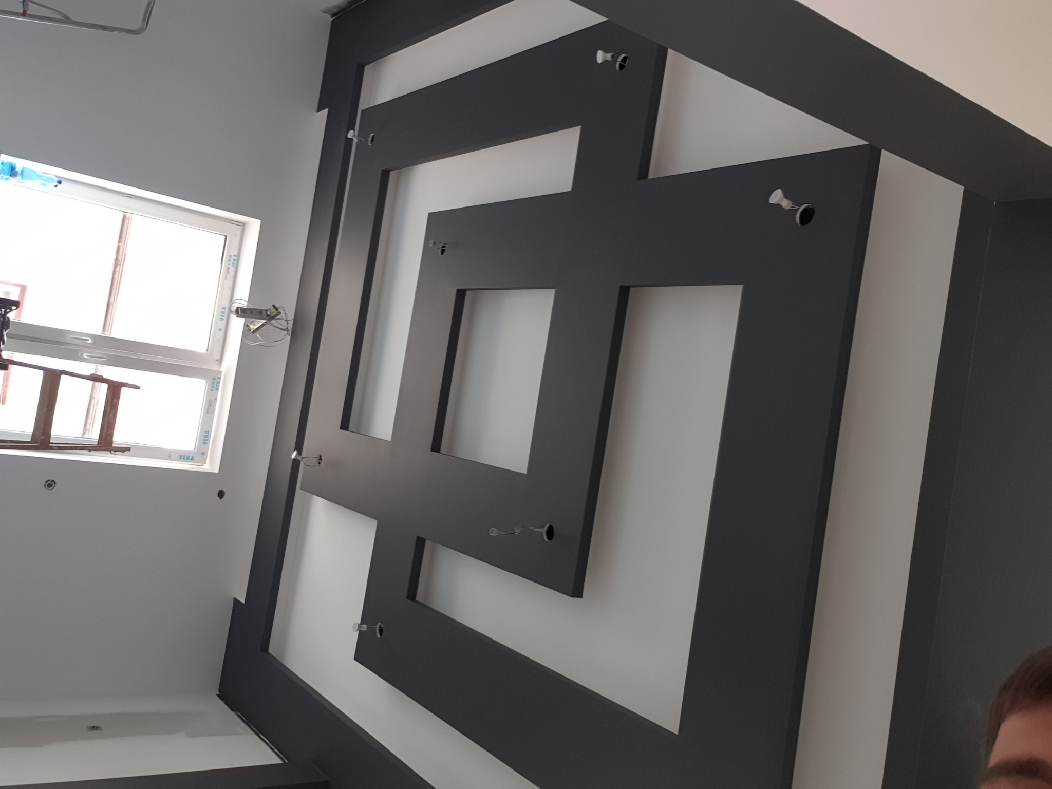 Geometryczny wzór z ciemnych listew na jasnej ścianie, widoczne otwory montażowe, fragment okna w tle