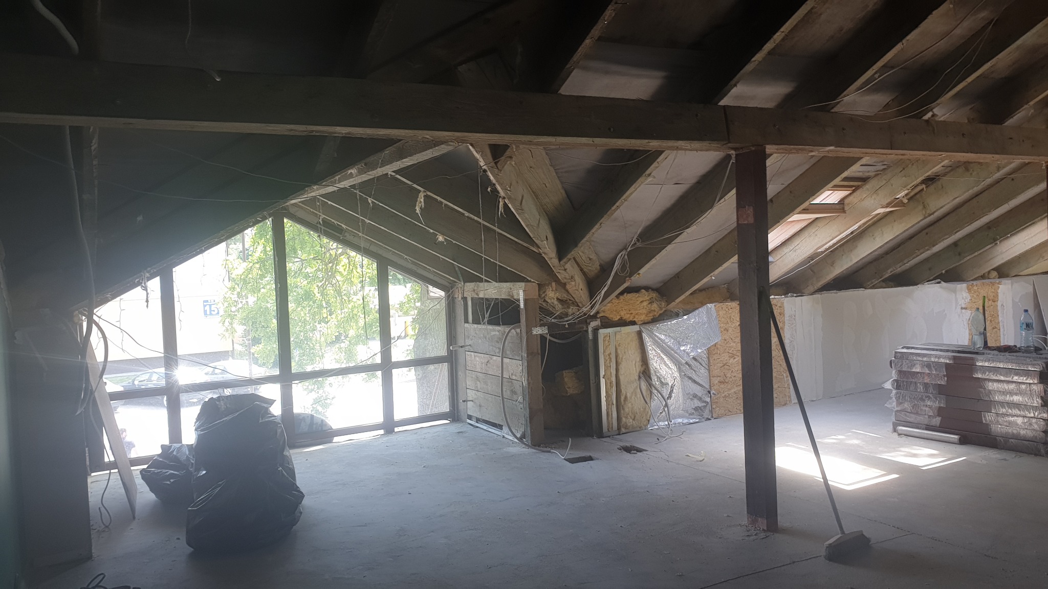 Poddasze w trakcie remontu: widoczne drewniane belki konstrukcyjne, częściowo ocieplone ściany, okno dachowe i dwa czarne worki na śmieci w rogu pomieszczenia.