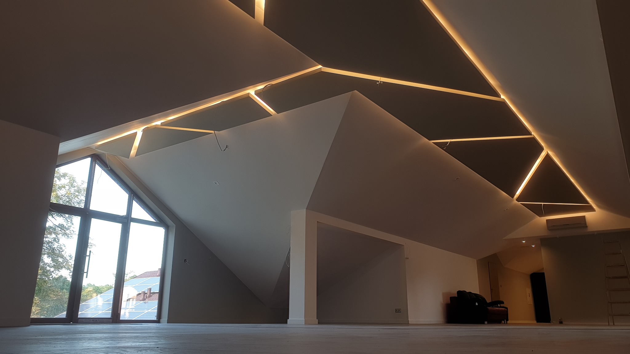 Poddasze po remoncie z oknem dachowym, białymi ścianami i sufitem z podświetlanymi panelami w geometryczne wzory.