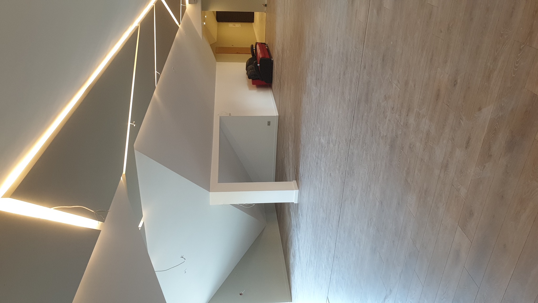 Nowoczesne wnętrze z podświetlanym sufitem o geometrycznych kształtach i panelami podłogowymi w odcieniach beżu; widoczne meble w oddali.