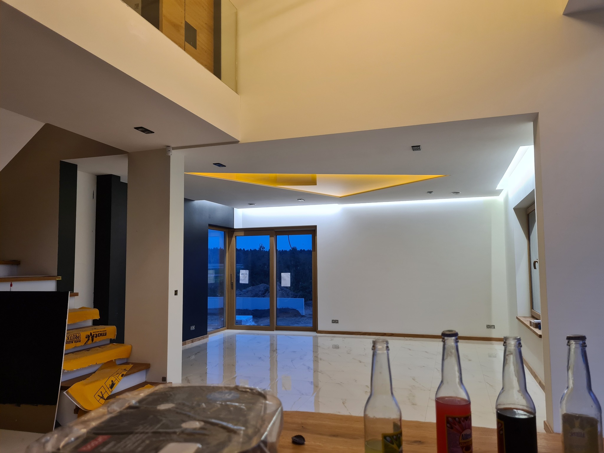Wnętrze domu w Lublinie w trakcie remontu, widoczne zabezpieczone schody folią budowlaną, błyszcząca marmurowa podłoga i butelki po napojach na pierwszym planie, sugerujące przerwę w pracy.