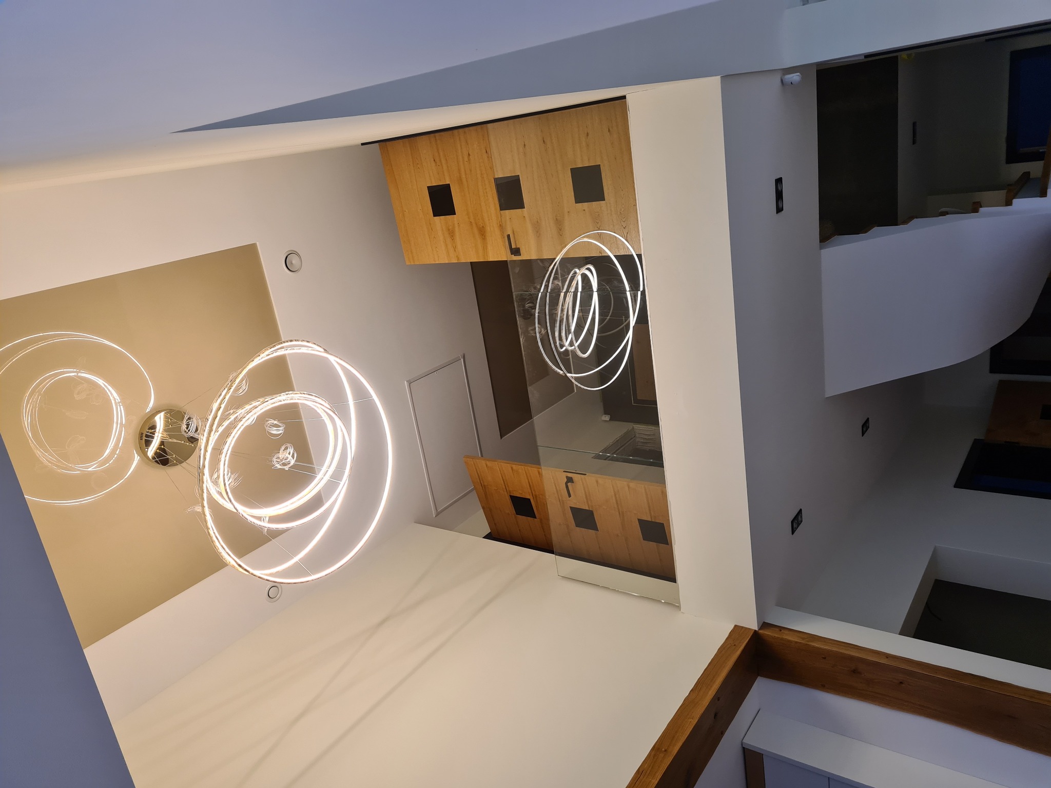 Nowoczesne wnętrze domu z widokiem na schody, drewniane elementy dekoracyjne i okrągłą lampę sufitową LED, uchwycone z perspektywy żabiej.