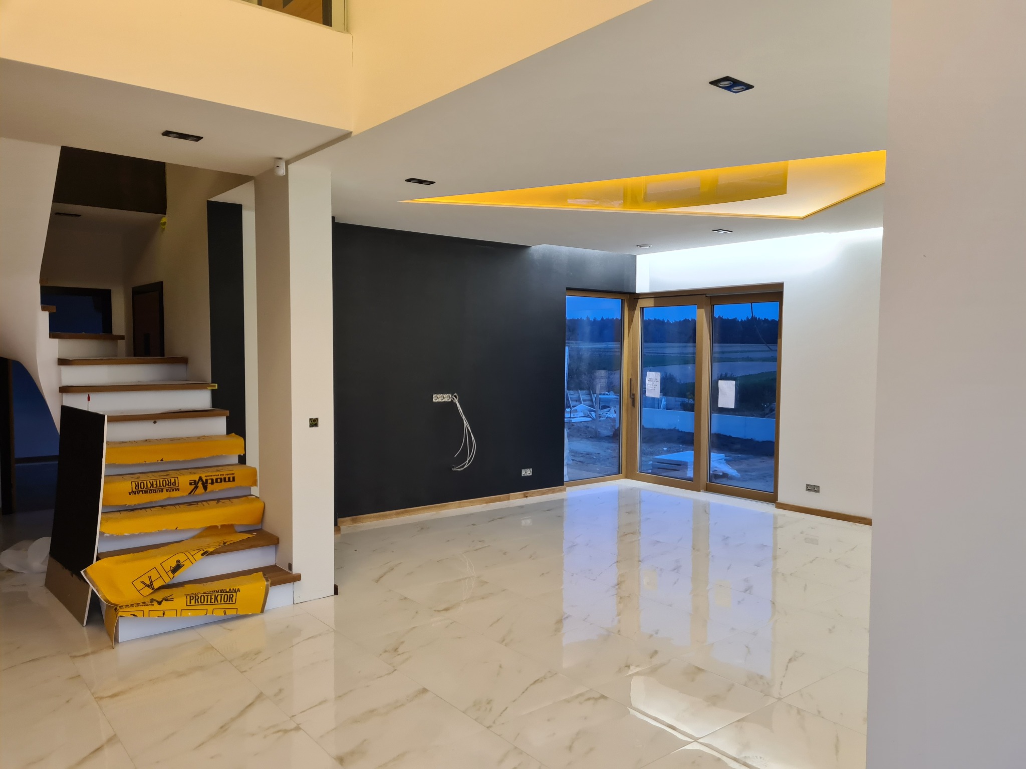 Wnętrze domu w trakcie wykańczania: marmurowe płytki podłogowe, zabezpieczone schody z drewnianymi stopniami, czarna ściana z wystającymi kablami, oświetlenie LED na suficie, duże okno tarasowe.